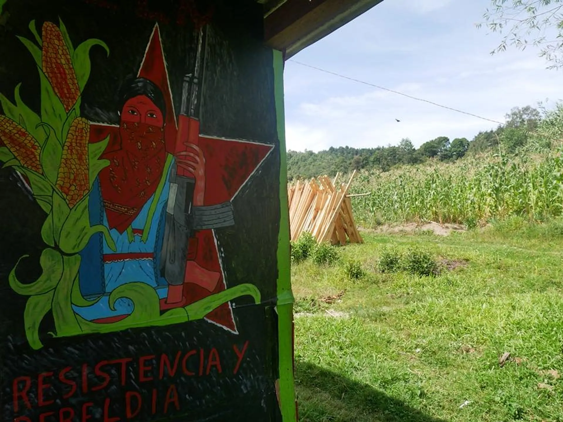 A mural depicting a masked figure with corn and a rifle, set against a lush cornfield in a rural area of Chiapas, Mexico.