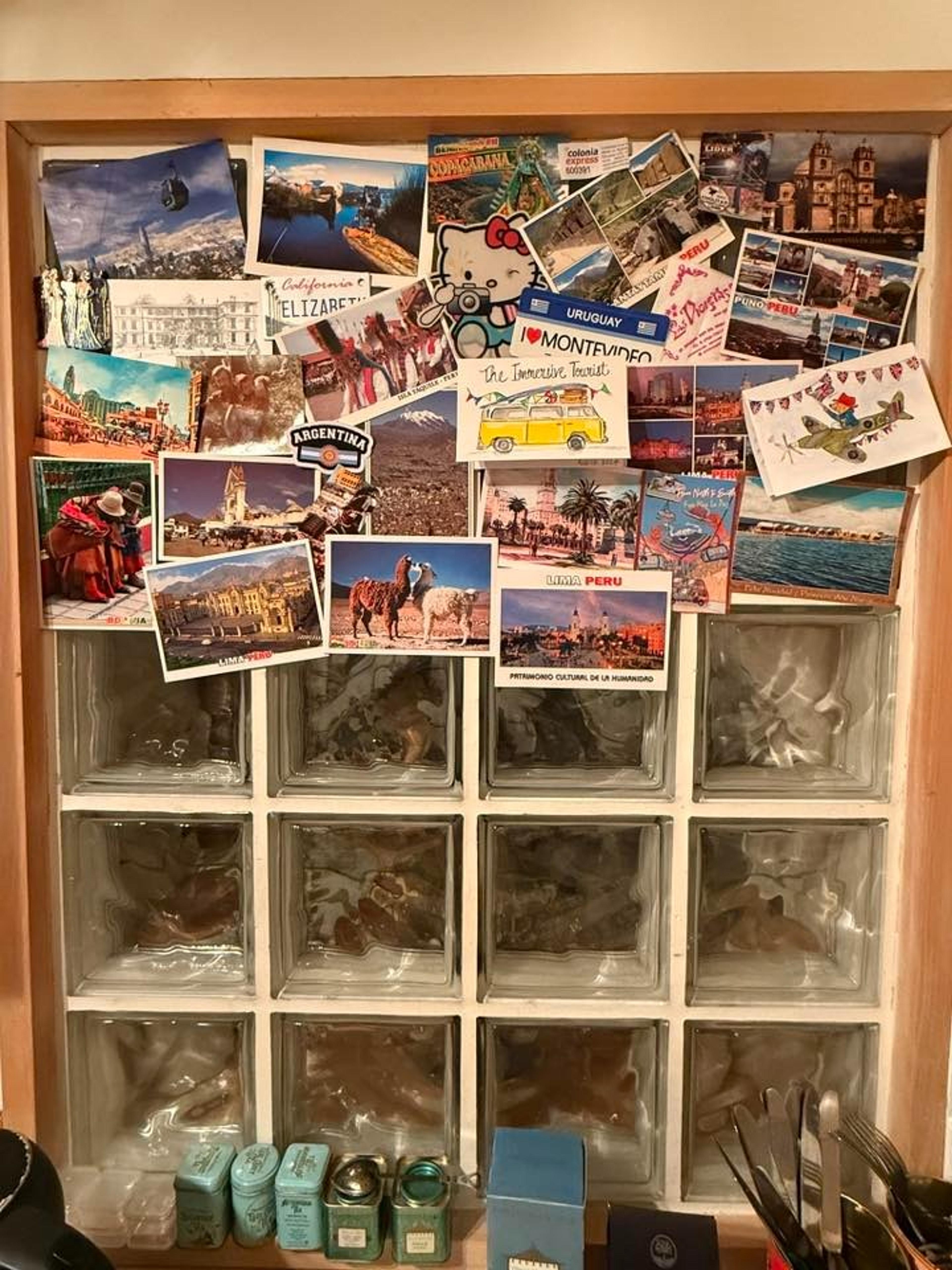 A collage of postcards from various global destinations decorates a glass-brick wall in a cozy indoor setting.