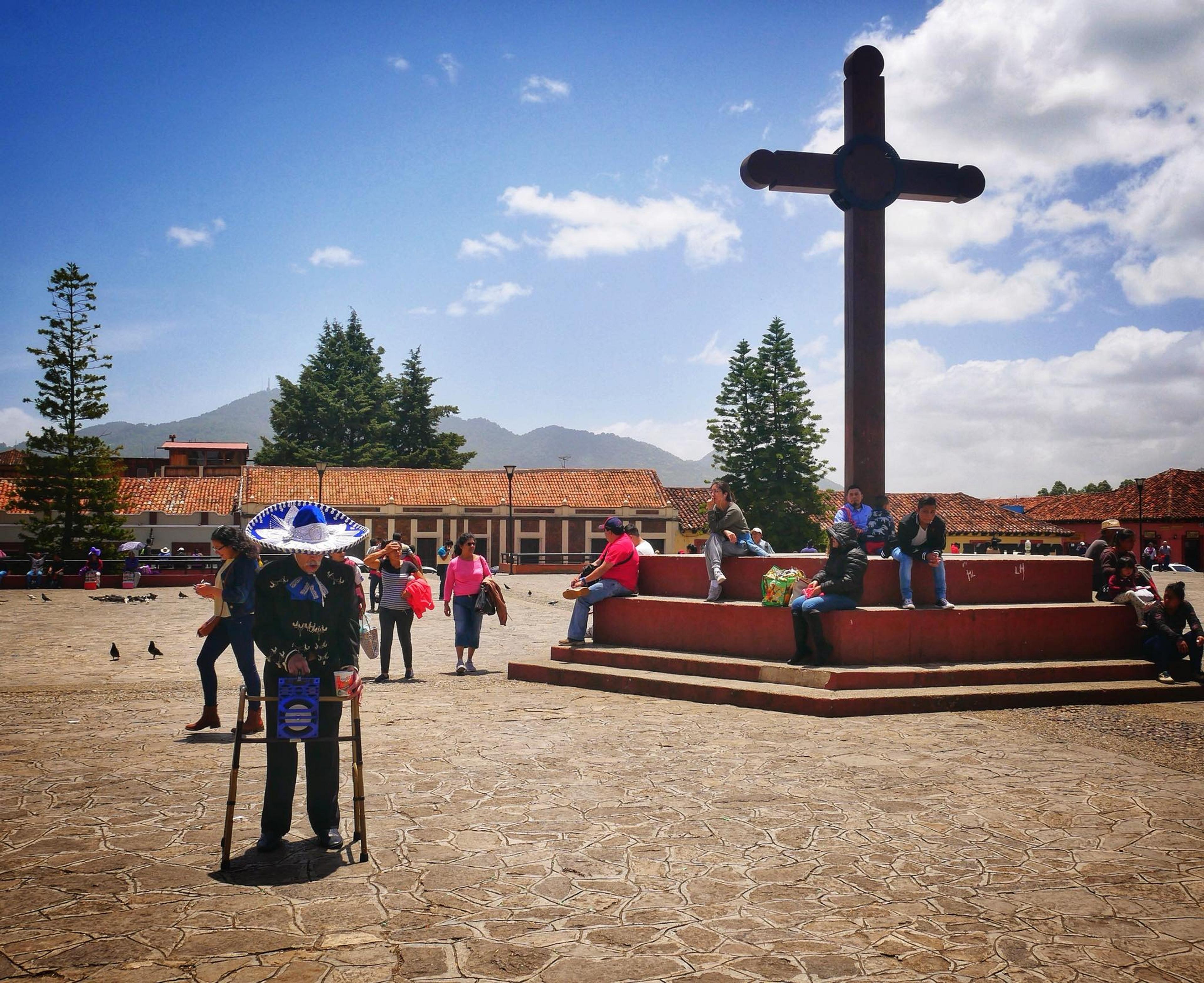 A lively scene in San Cristóbal de las Casas, Mexico, features people gathered around a large wooden cross in a central plaza against a backdrop of mountains and colonial-style buildings.
