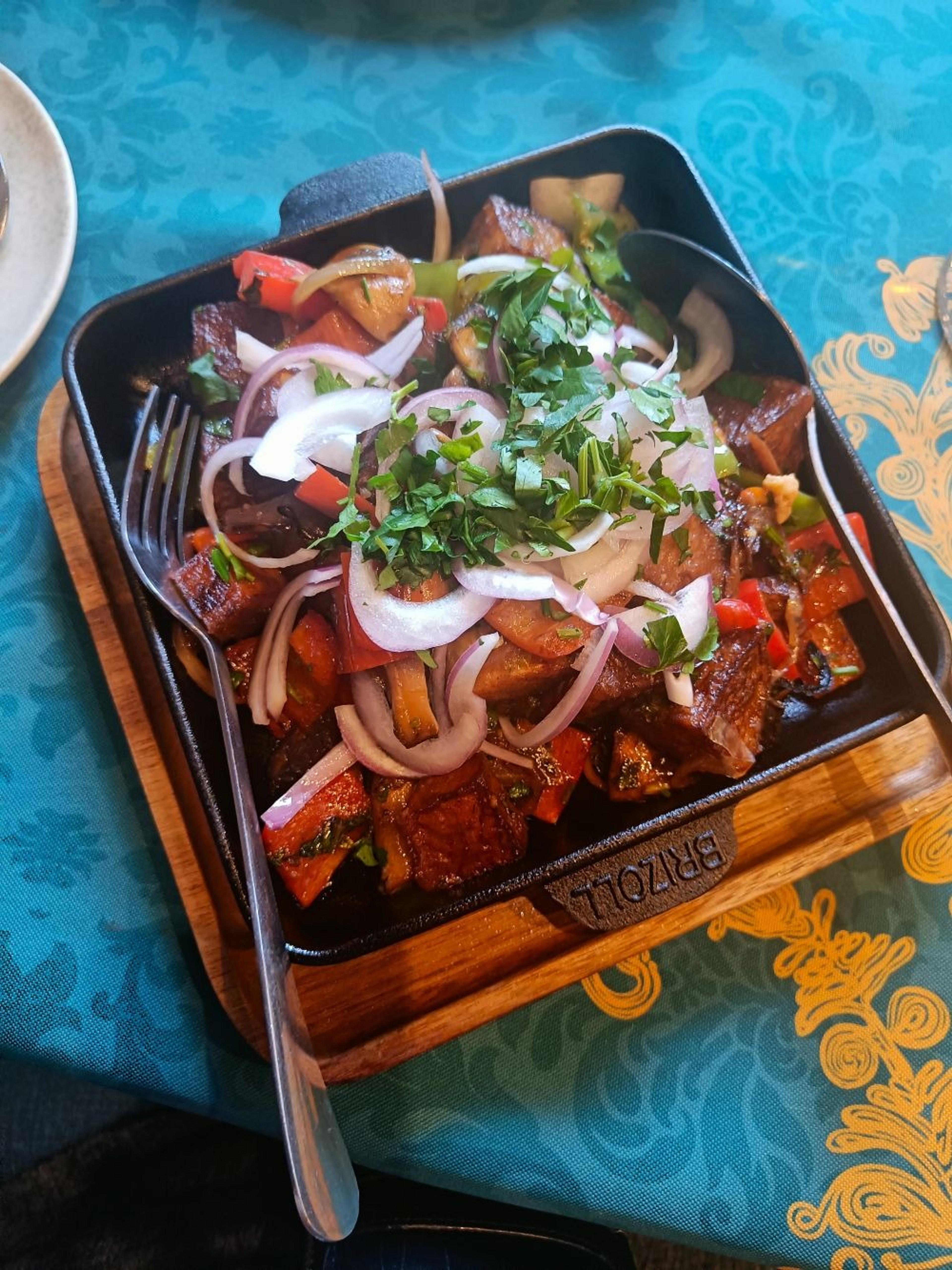 A sizzling skillet of roasted vegetables adorned with fresh herbs and onions sits on a turquoise-patterned tablecloth in Tbilisi