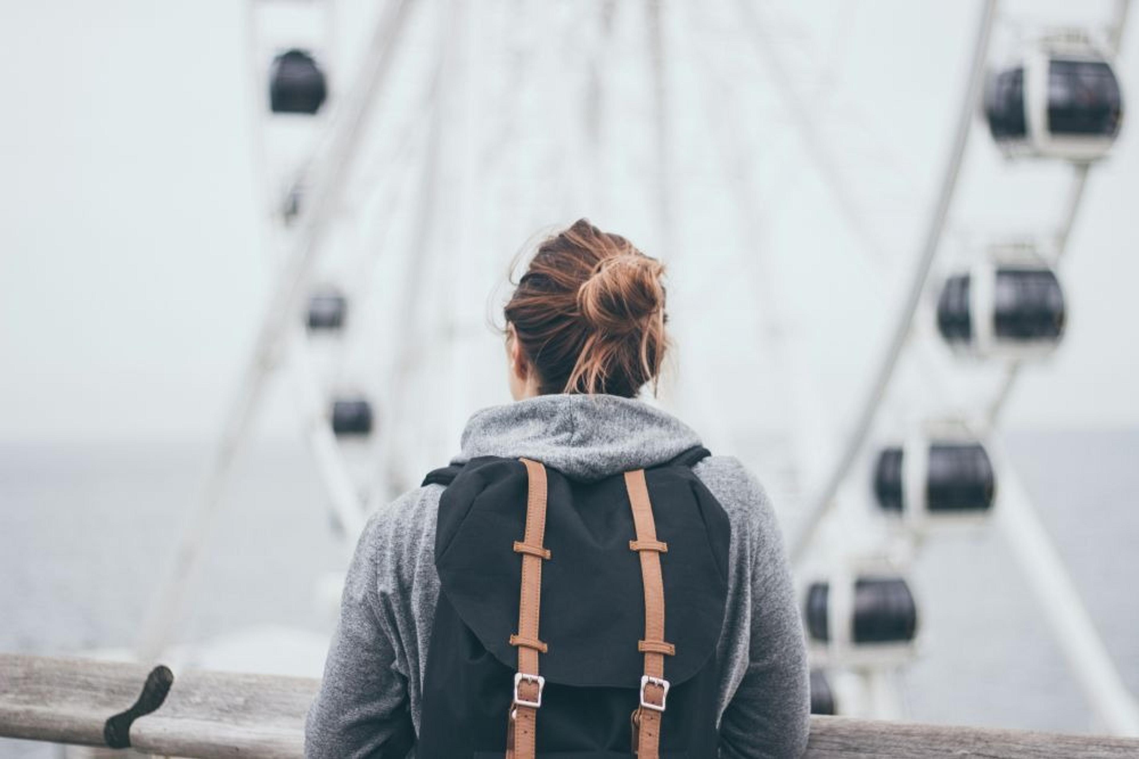 A person with a backpack stands near a large Ferris wheel by what appears to be a seaside location.