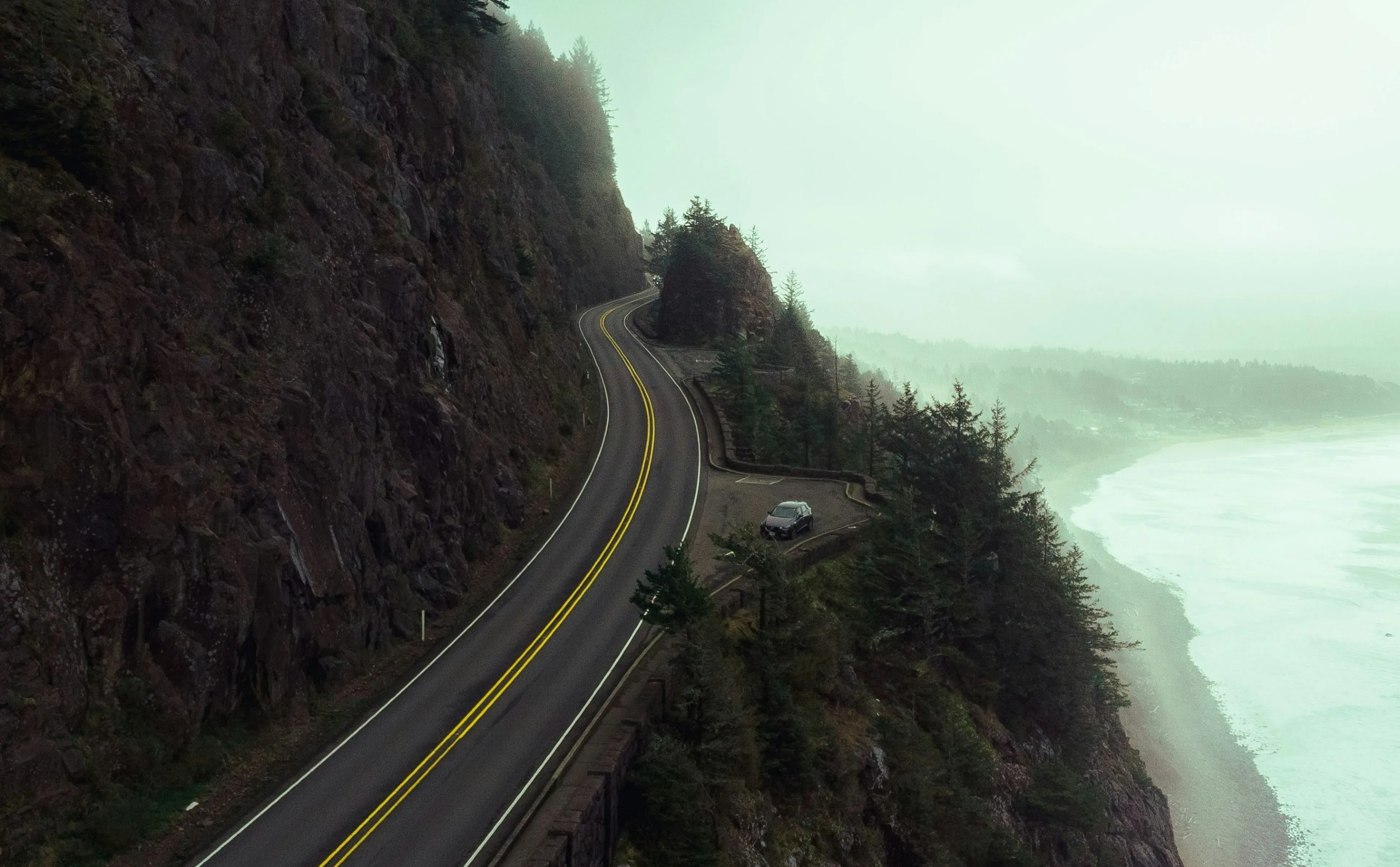 A coastal highway winds along a cliffside in Oregon, overlooking a misty ocean and dense forest.