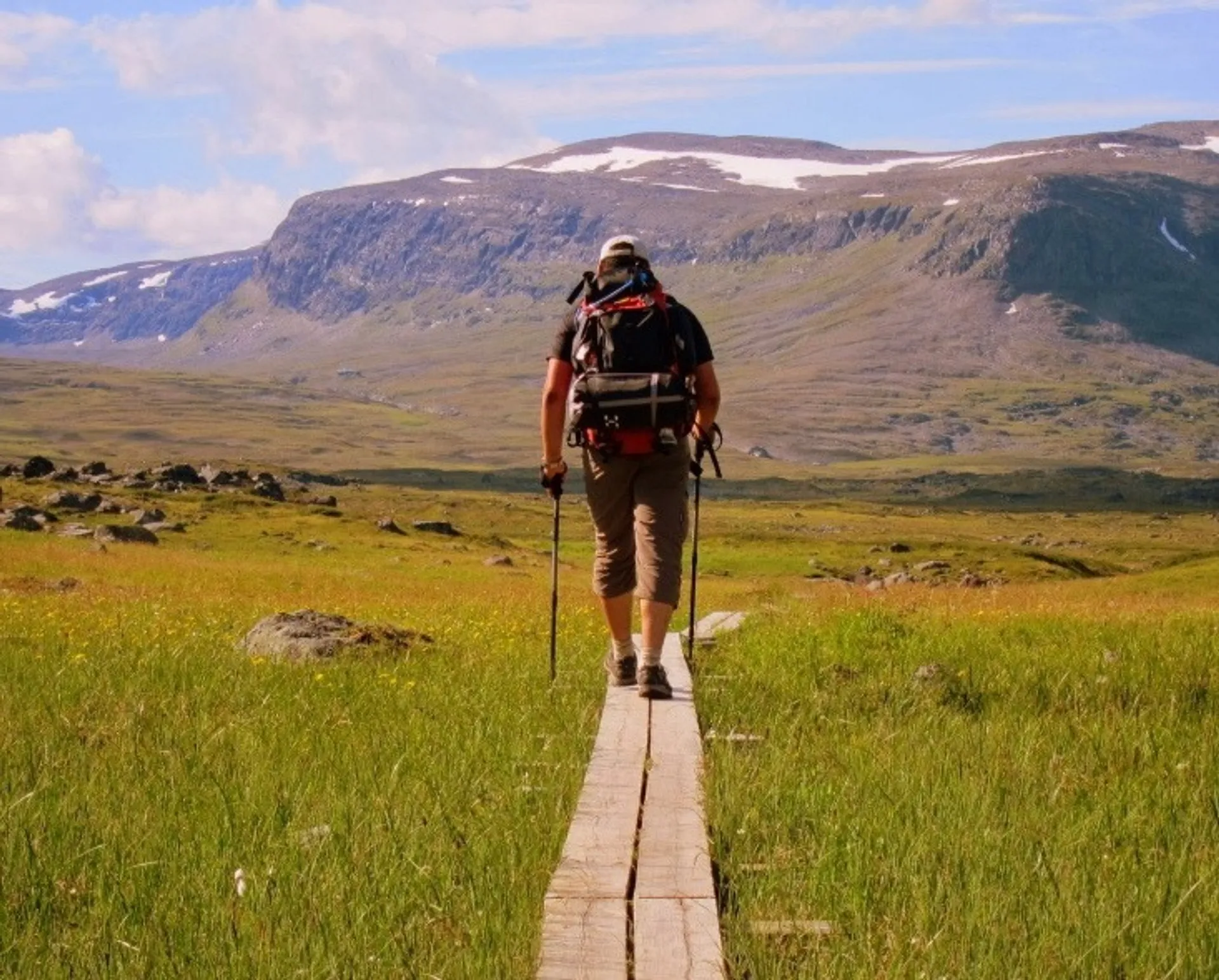 A hiker with trekking poles walks along a wooden path in the Kungsleden trail in Sweden, surrounded by grassy fields and mountain scenery.