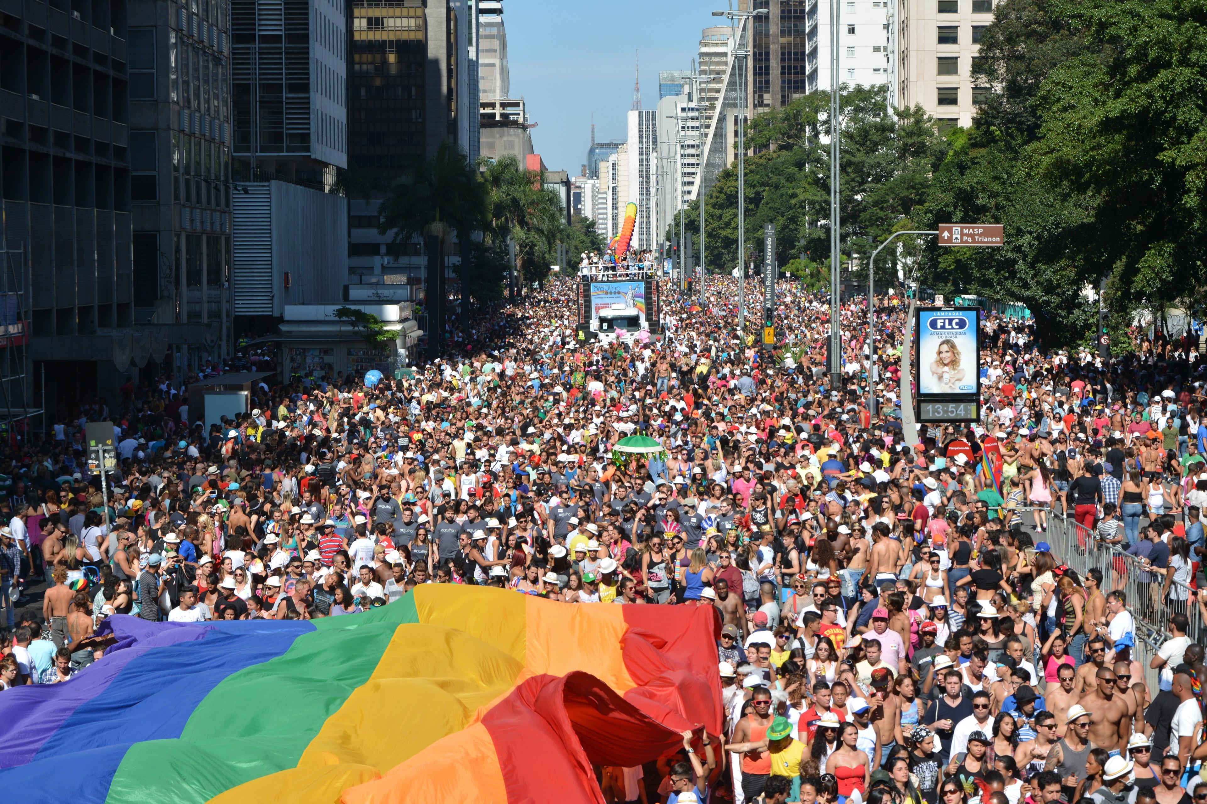 A vibrant and massive crowd gathers for a festival or parade on Avenida Paulista in São Paulo, featuring a large rainbow flag. 