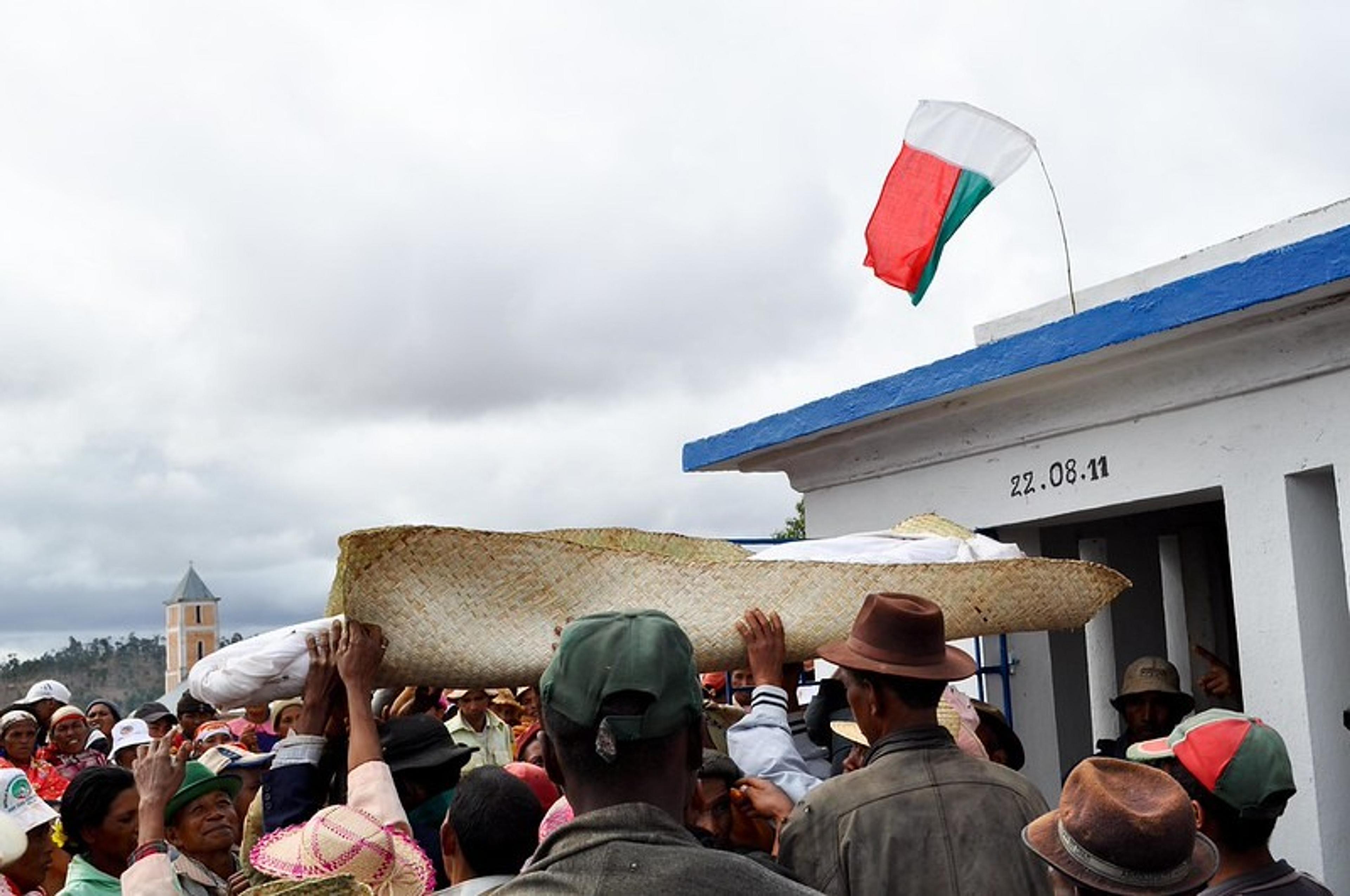 A crowd participates in a traditional burial ceremony in Madagascar, marked by the national flag and the carrying of a wrapped body.