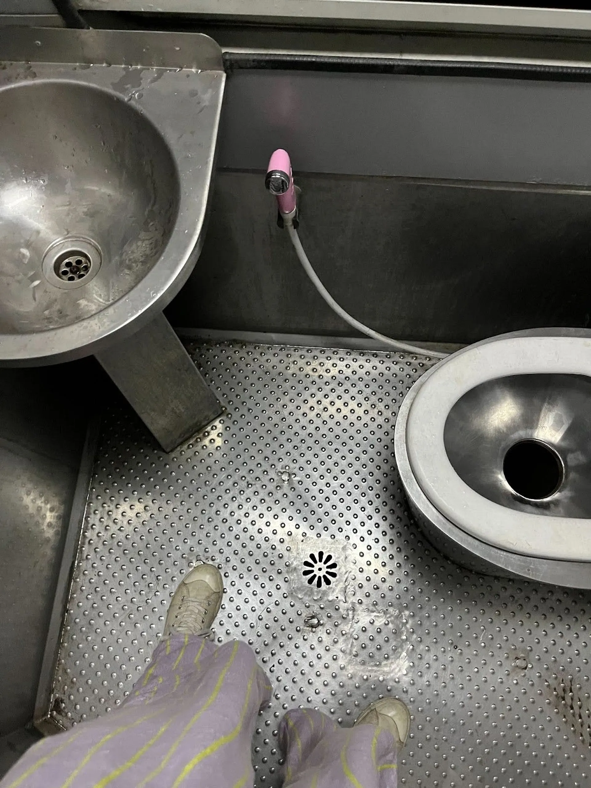 A stainless steel bathroom in a train compartment with a toilet, sink, and hose.
