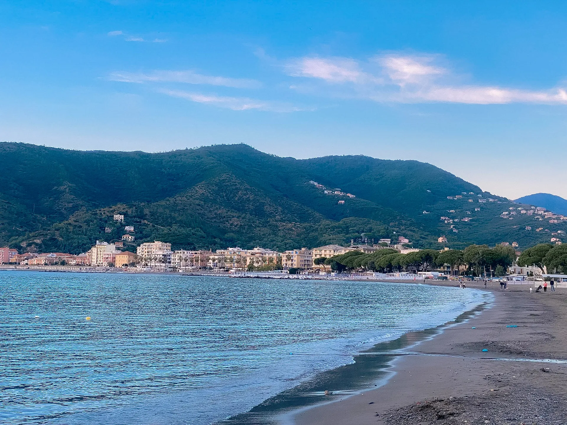 A peaceful beach scene unfolds along the shoreline with lush green hills and charming seaside buildings.