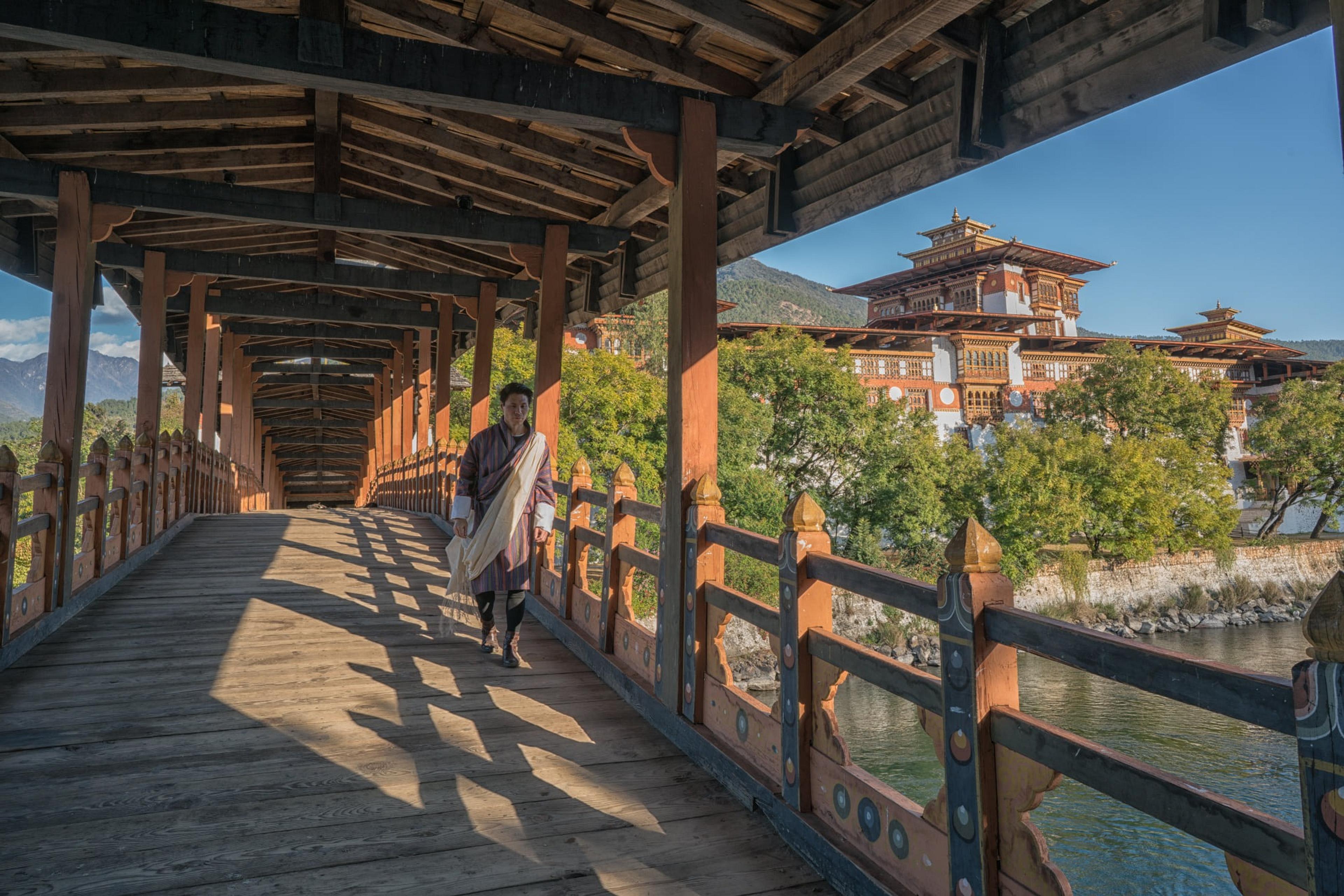 A traditional wooden bridge leads to the majestic Punakha Dzong in Bhutan, surrounded by lush greenery and set against a mountainous backdrop.