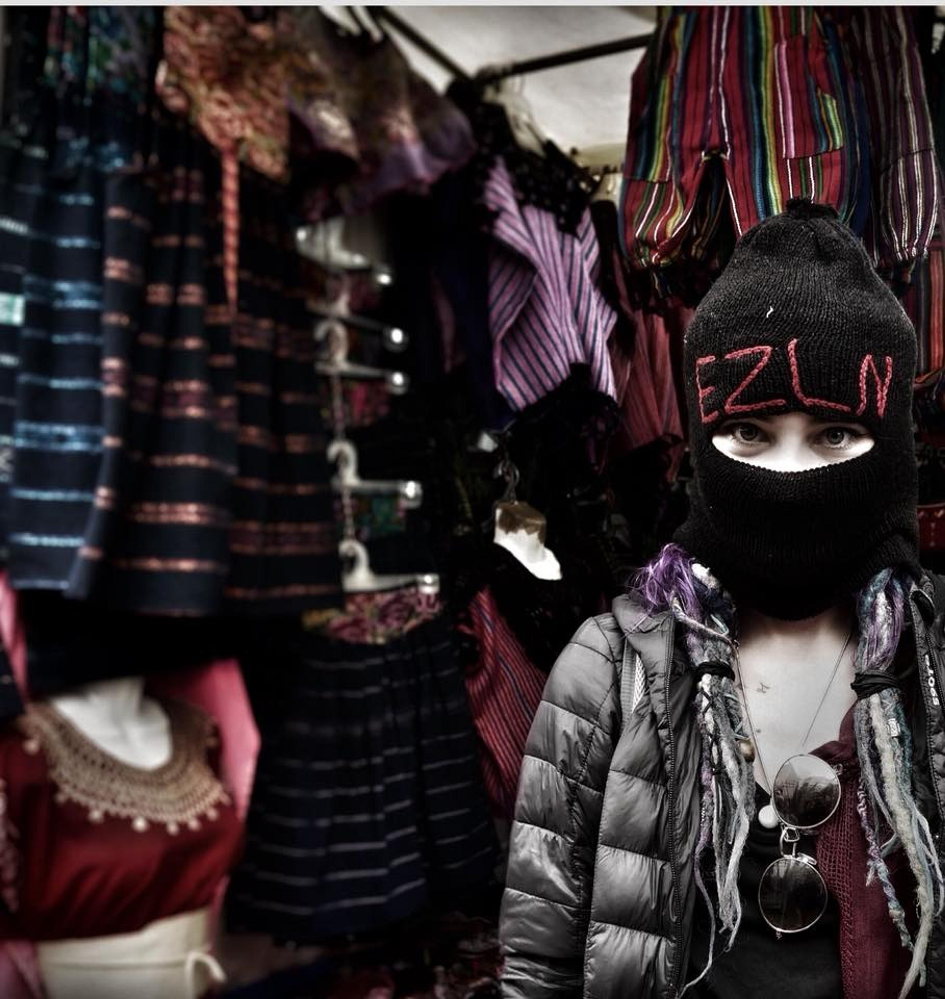 A person wearing an "EZLN" balaclava stands in front of a colorful textile stall in southern Mexico.