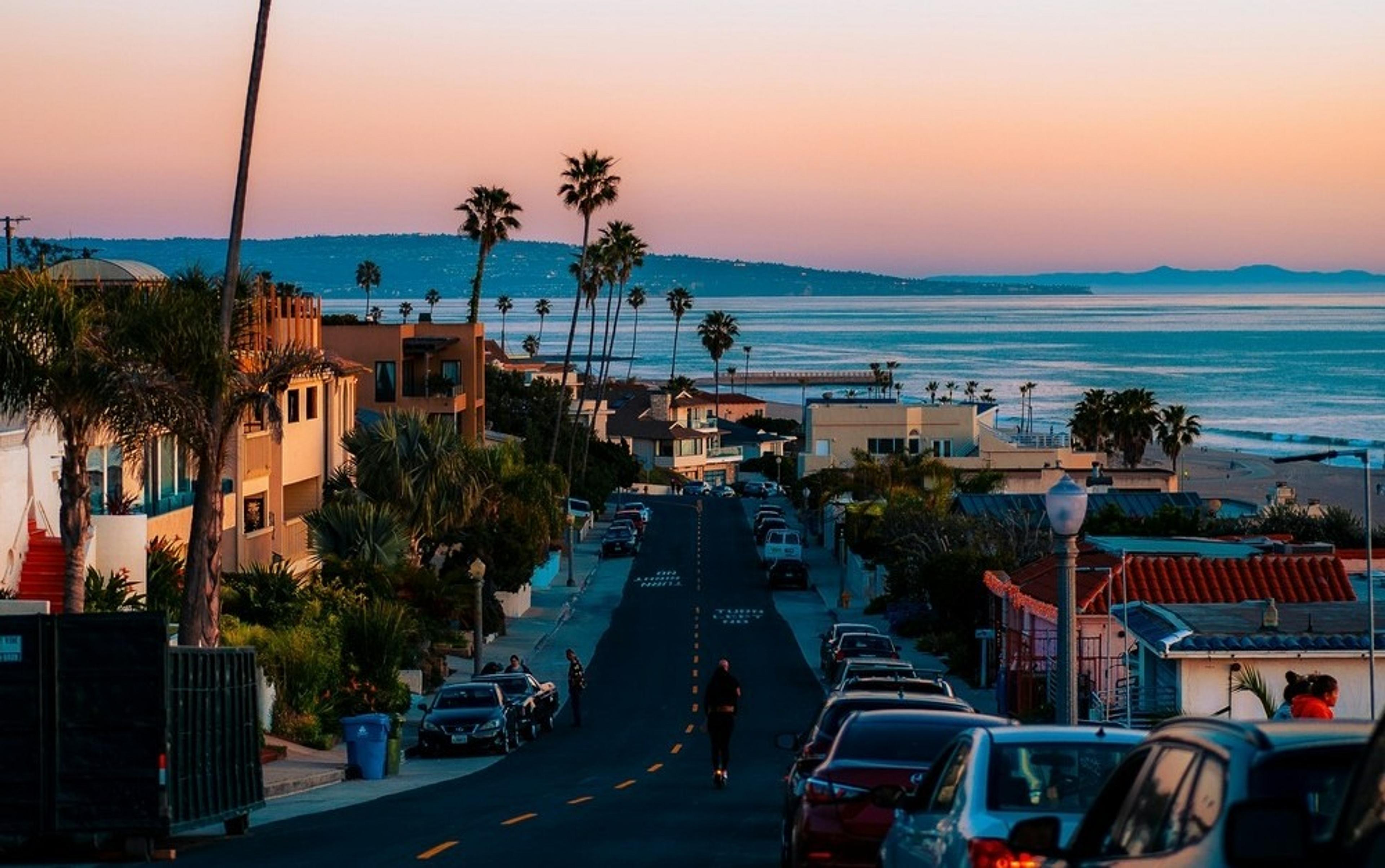 A coastal street in Manhattan Beach, California, gently slopes towards the ocean at sunset with palm trees lining the view.