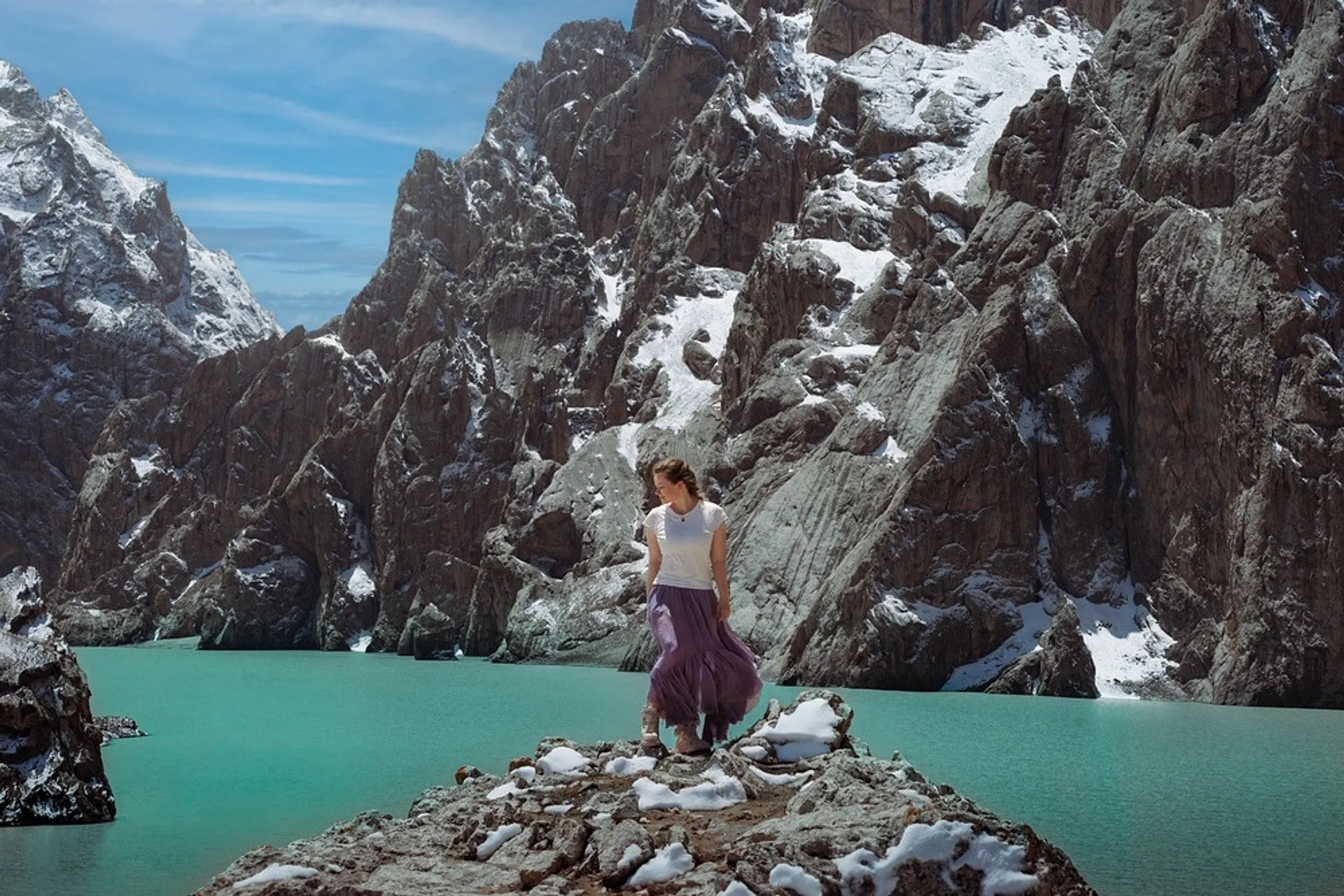 Flight Finder Kristi stands at Kel-Suu, a turquoise mountain lake, surrounded by rugged, snow-dusted cliffs.