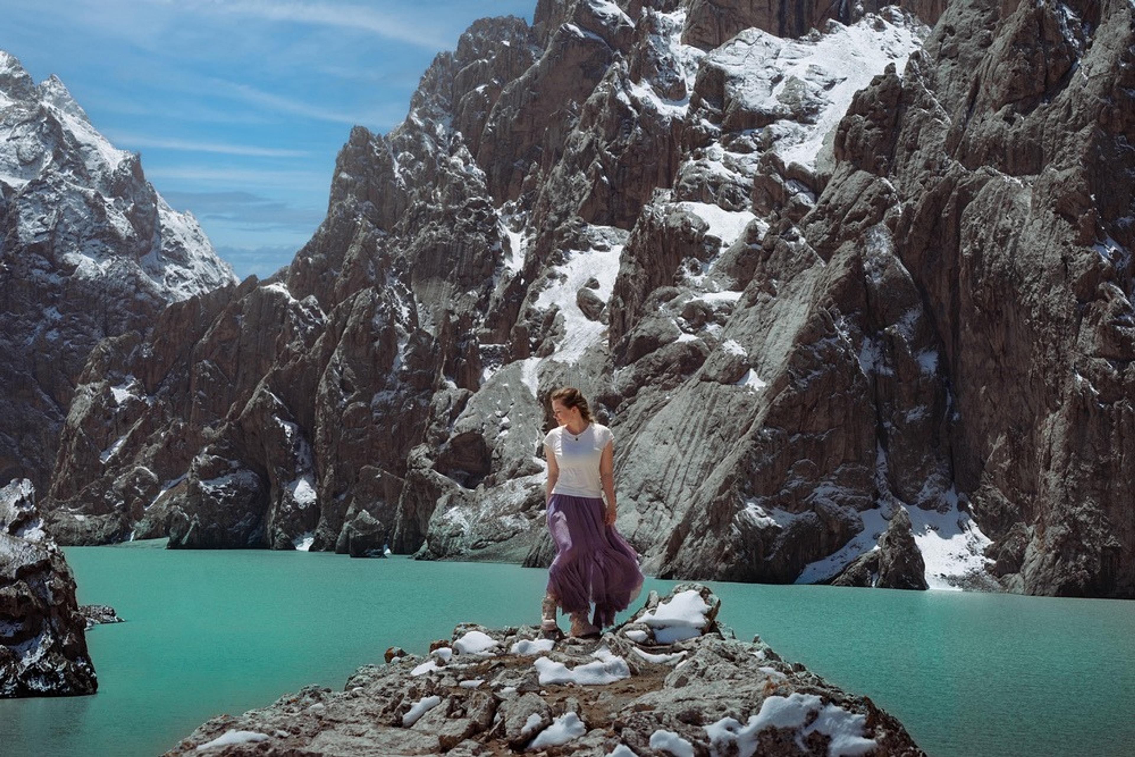 Flight Finder Kristi stands at Kel-Suu, a turquoise mountain lake, surrounded by rugged, snow-dusted cliffs.
