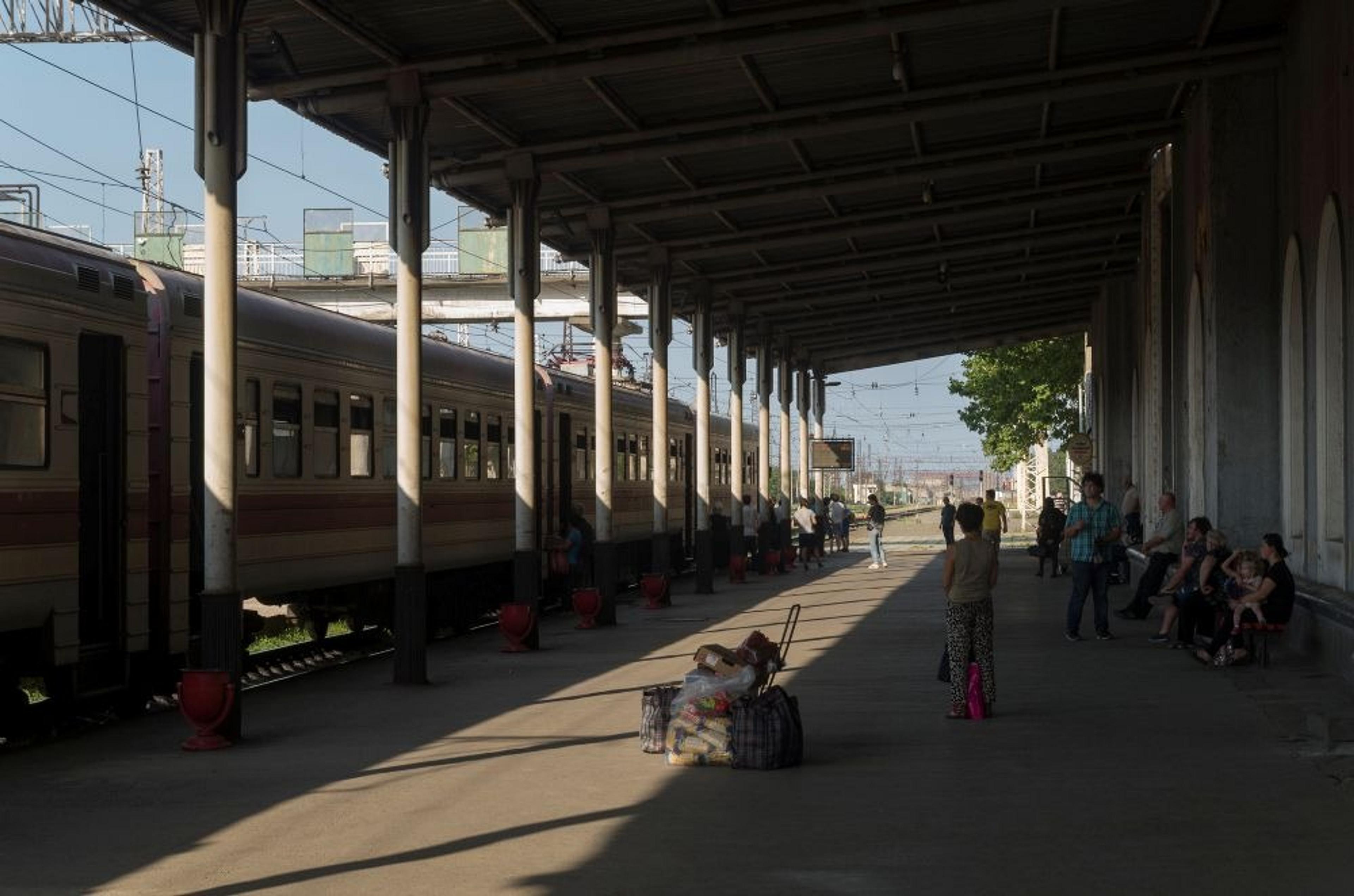 A train station platform in Vladikavkaz, Russia, hosts passengers waiting by a stationary train under a large metal canopy.