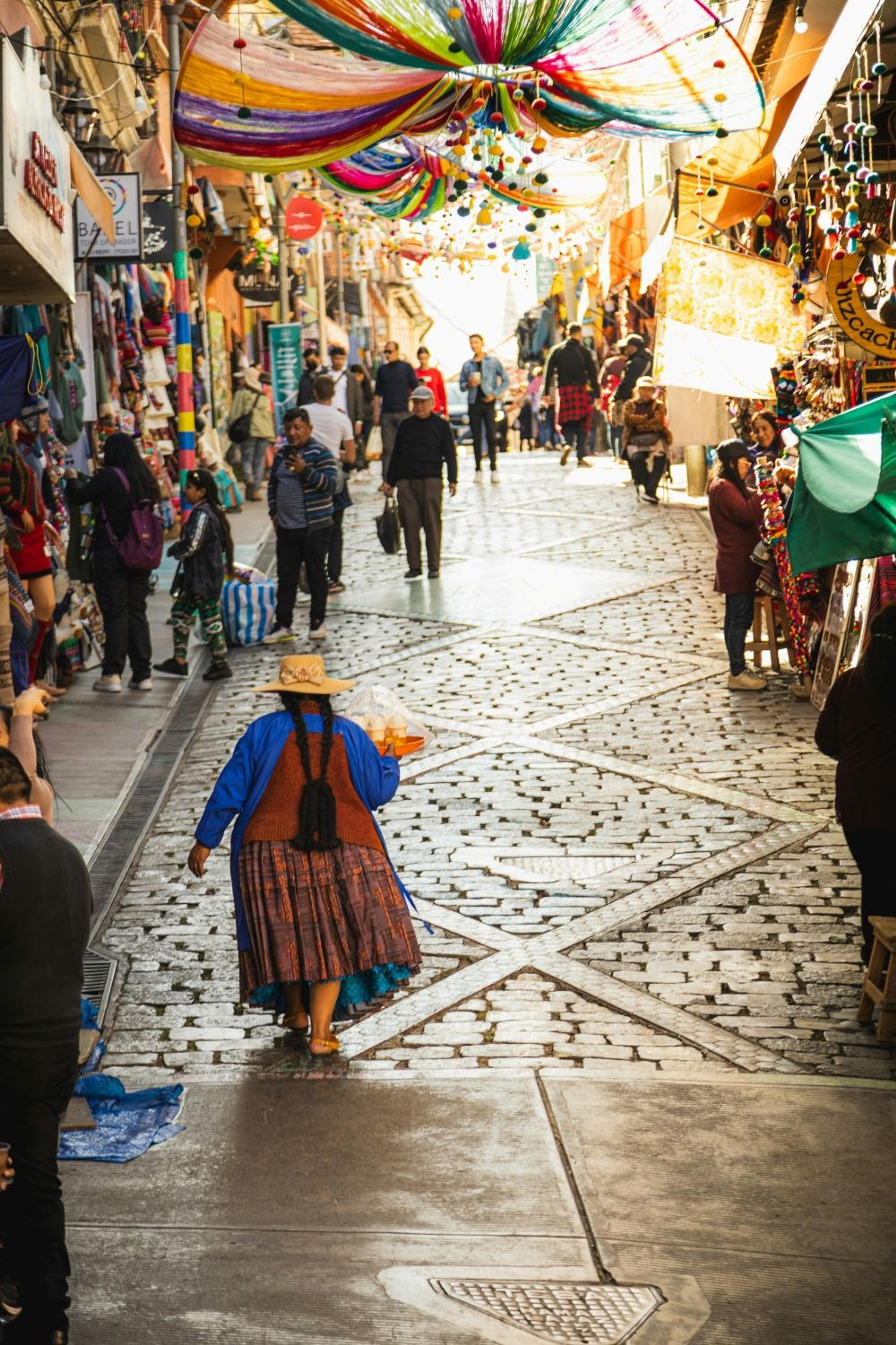 A vibrant, bustling street market in La Paz, Bolivia is adorned with colorful canopy decorations and busy with shoppers.