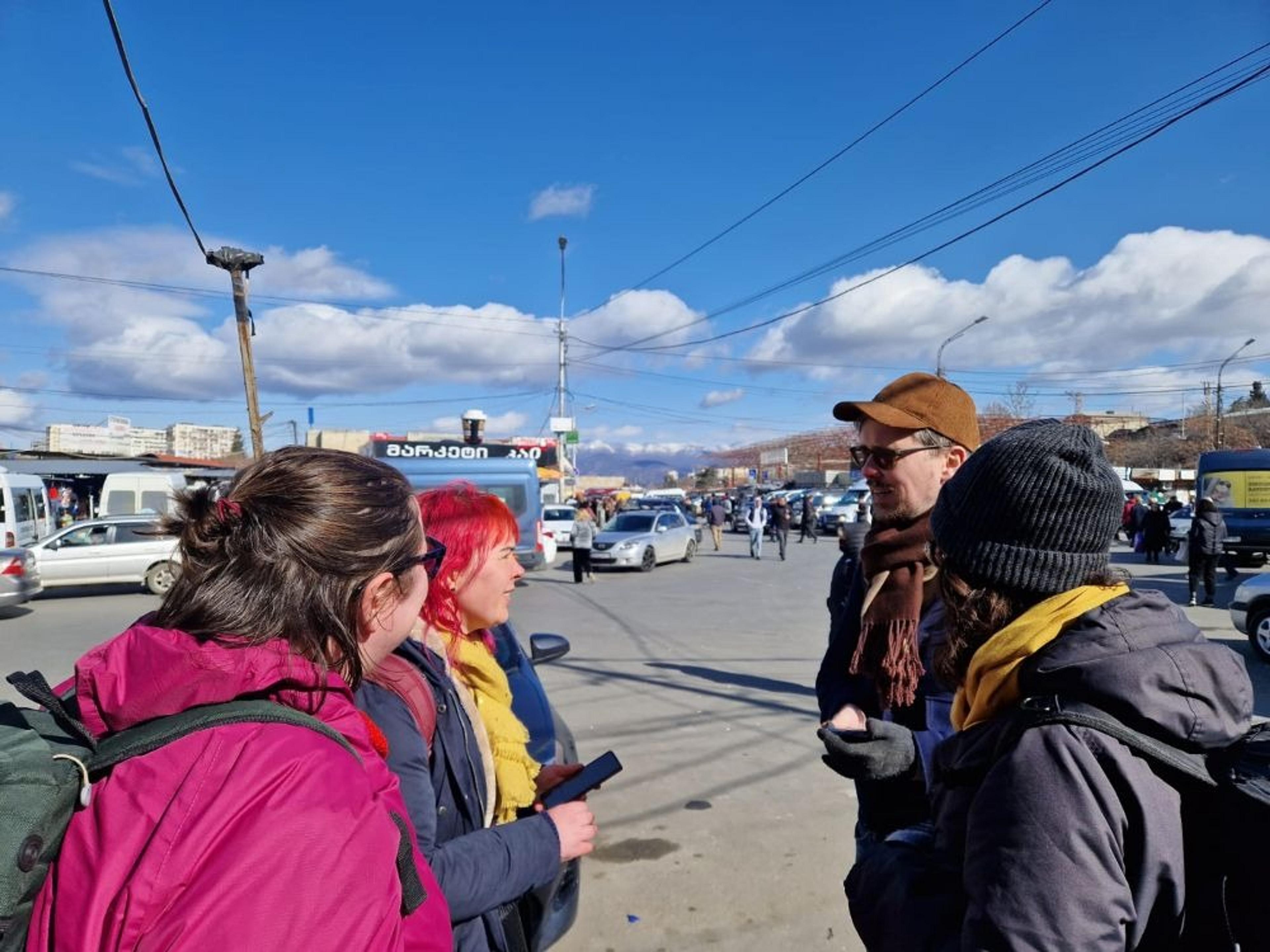 A group of people converse on a bustling street near a market in Tbilisi, Georgia, beneath a bright blue sky.