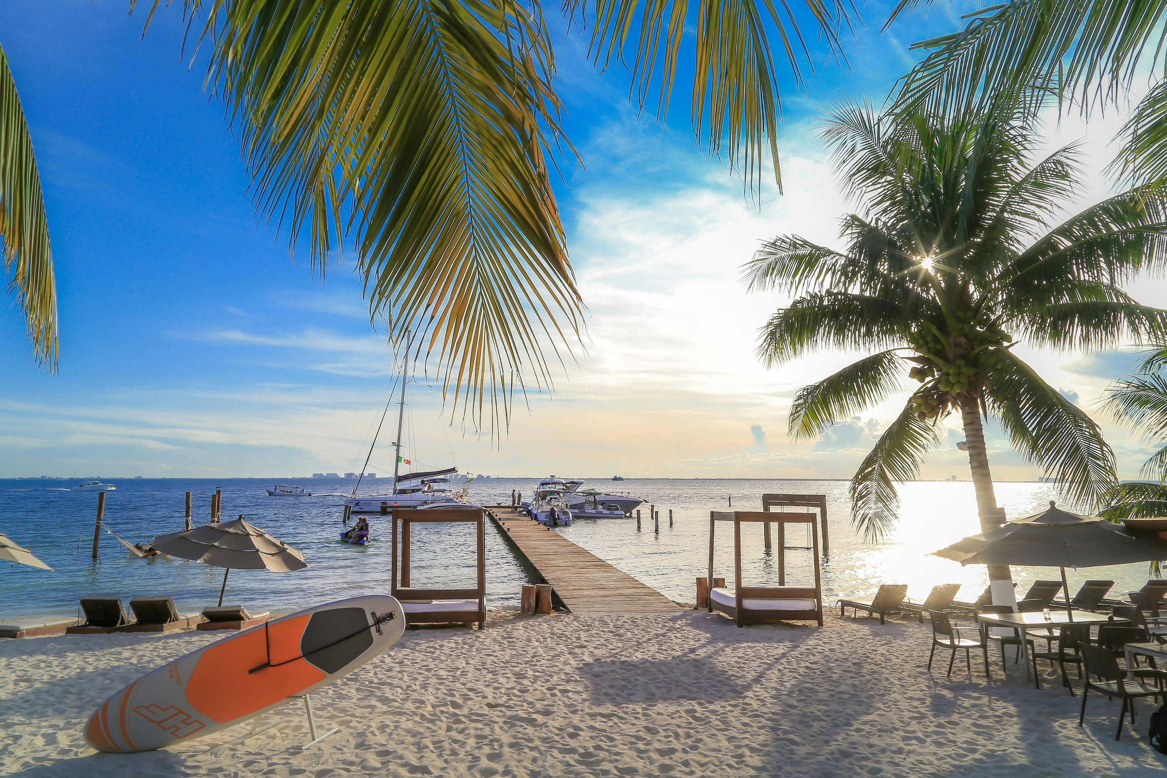 A serene beach scene in Isla Mujeres, Mexico, features inviting palm trees, lounge chairs, and a pier extending into a tranquil sea under a bright blue sky.
