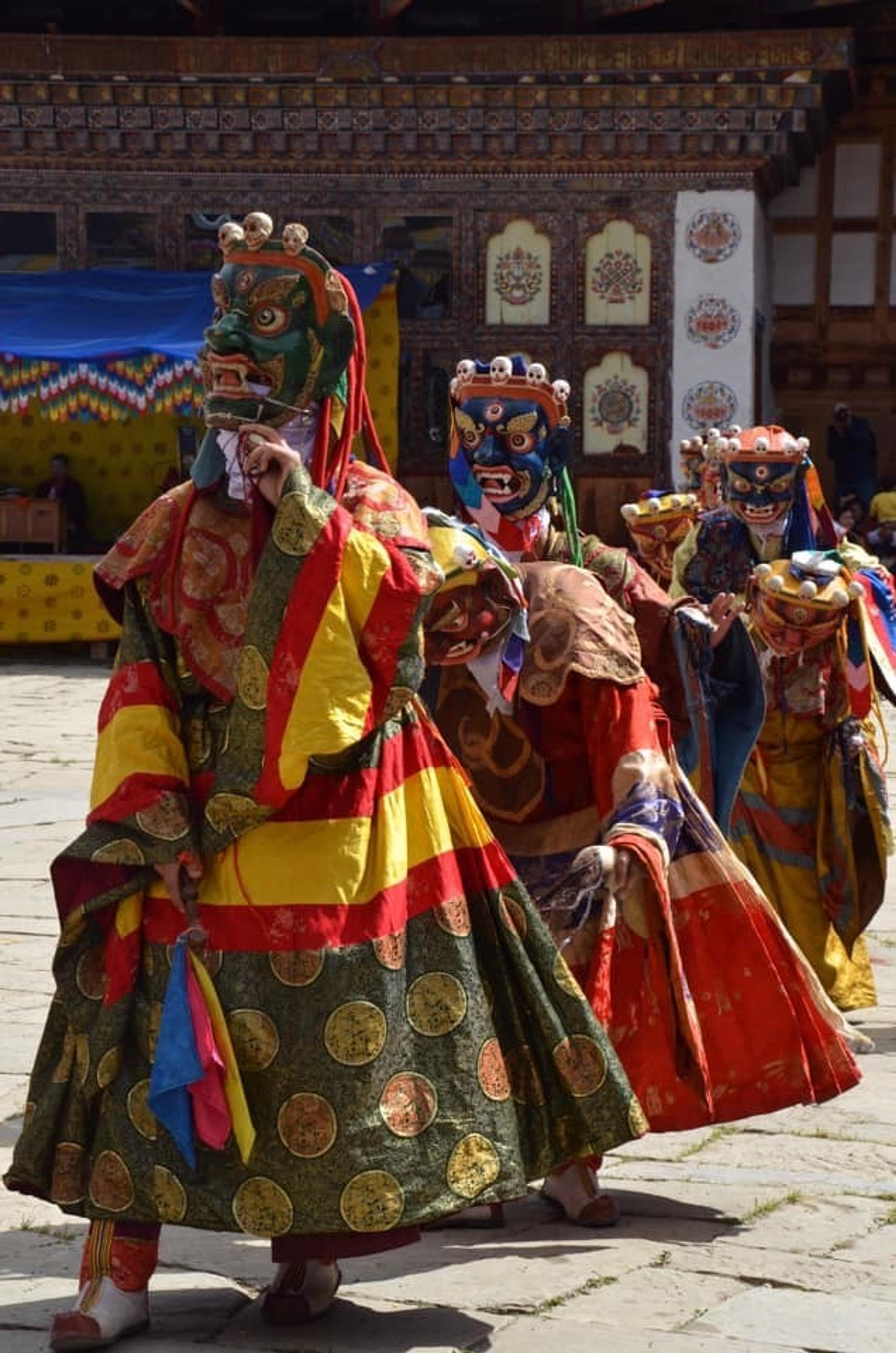 Performers wearing vibrant traditional masks and costumes participate in a festival at a dzong, likely in Bhutan.