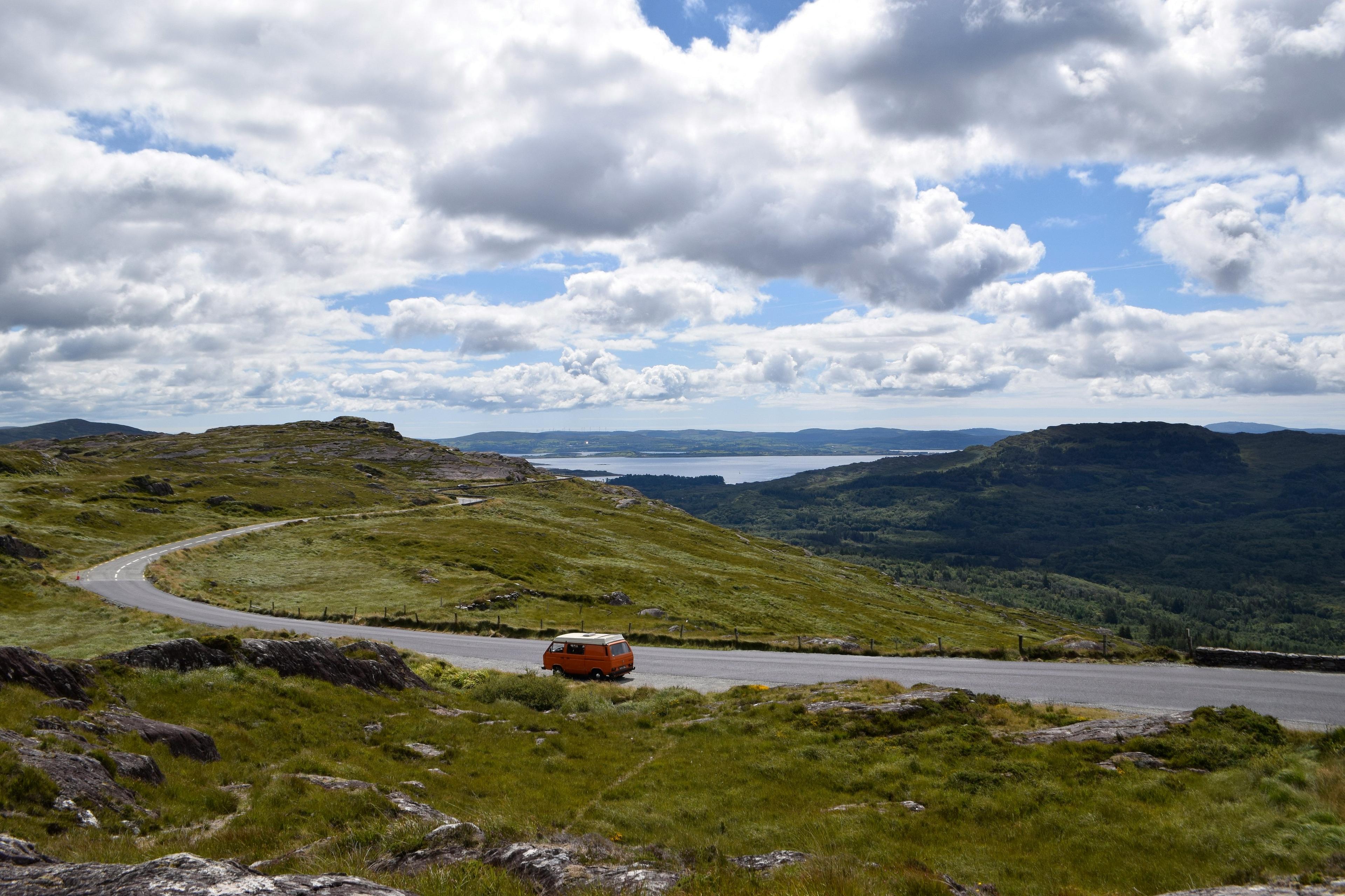 An orange van travels along a winding road in the scenic landscape owith lush greenery and distant hills under a partly cloudy sky.