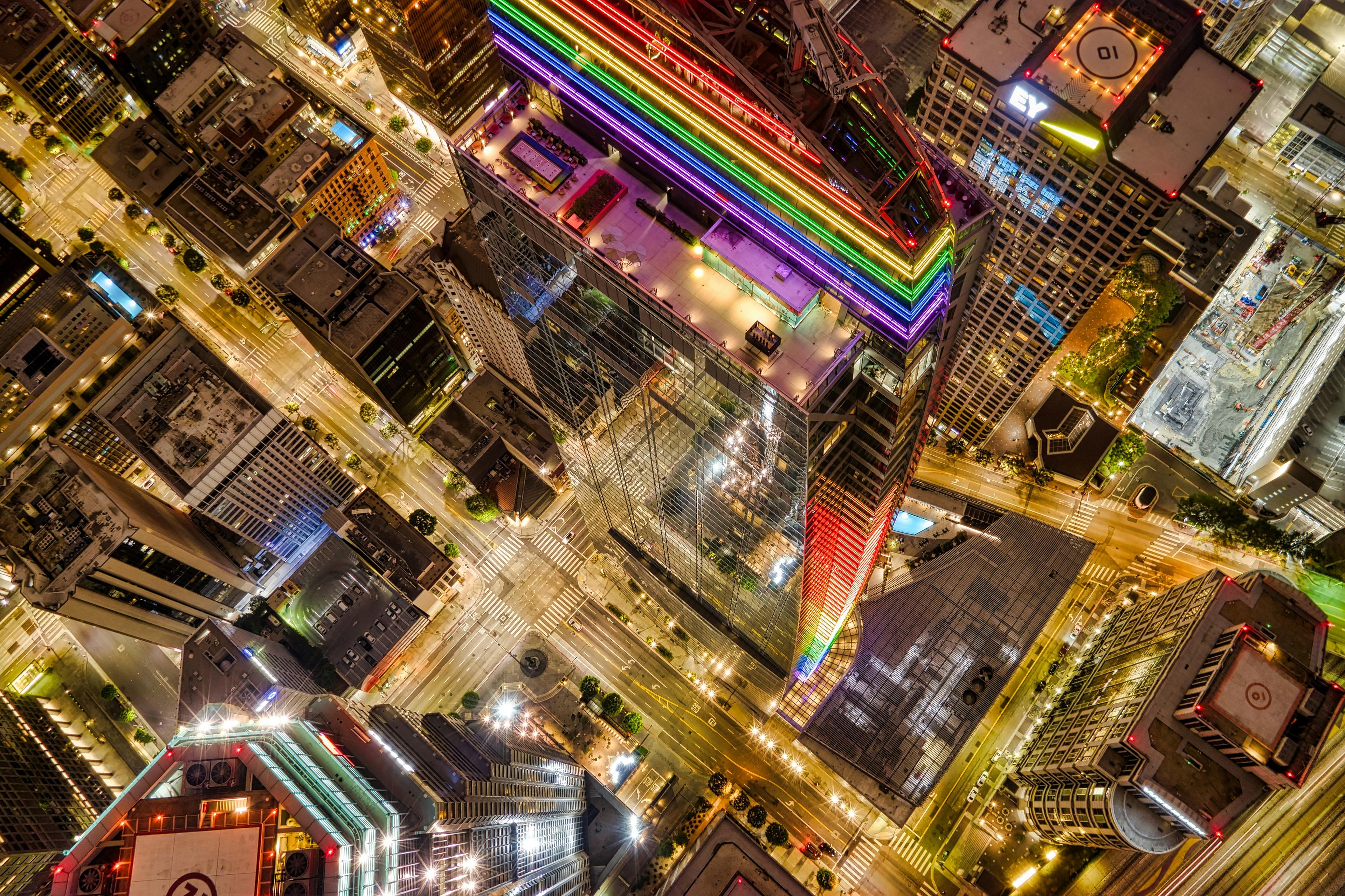 An aerial view of downtown Los Angeles at night features a building illuminated with rainbow lights.