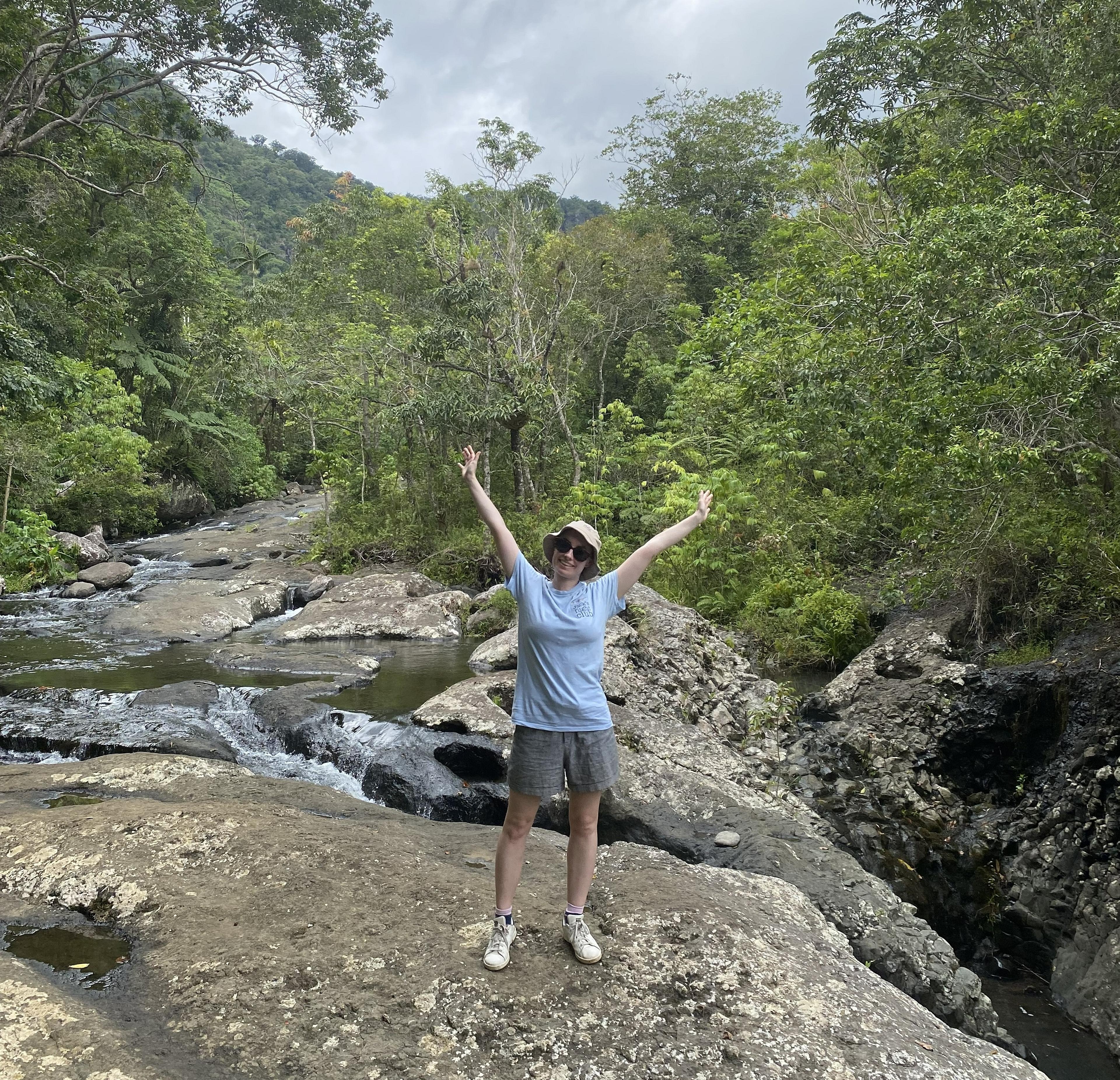 A person joyfully poses near a rocky stream surrounded by lush greenery in a tropical forest setting.