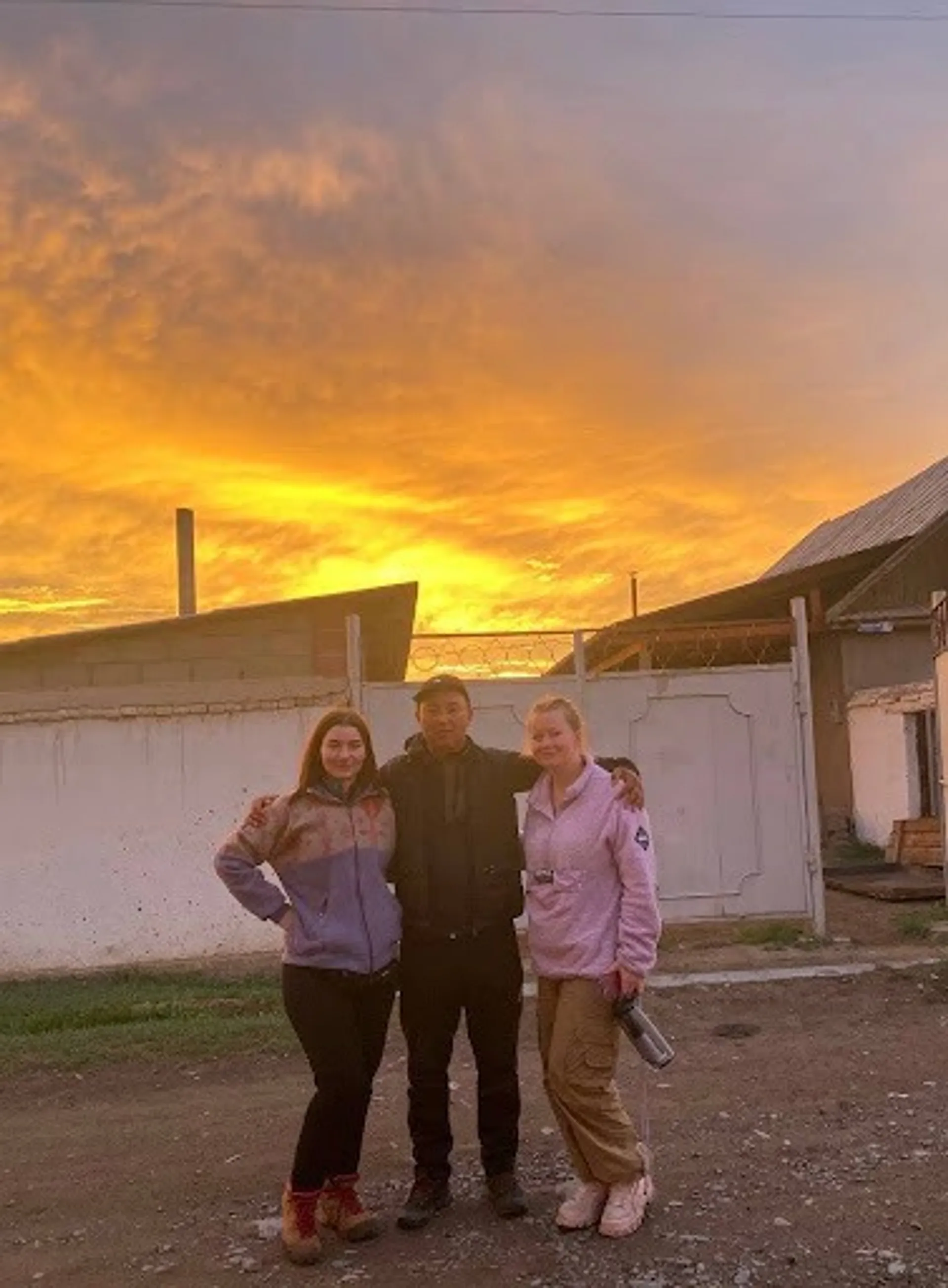 Three people stand together under a vibrant sunset in a rural area in Kyzart.