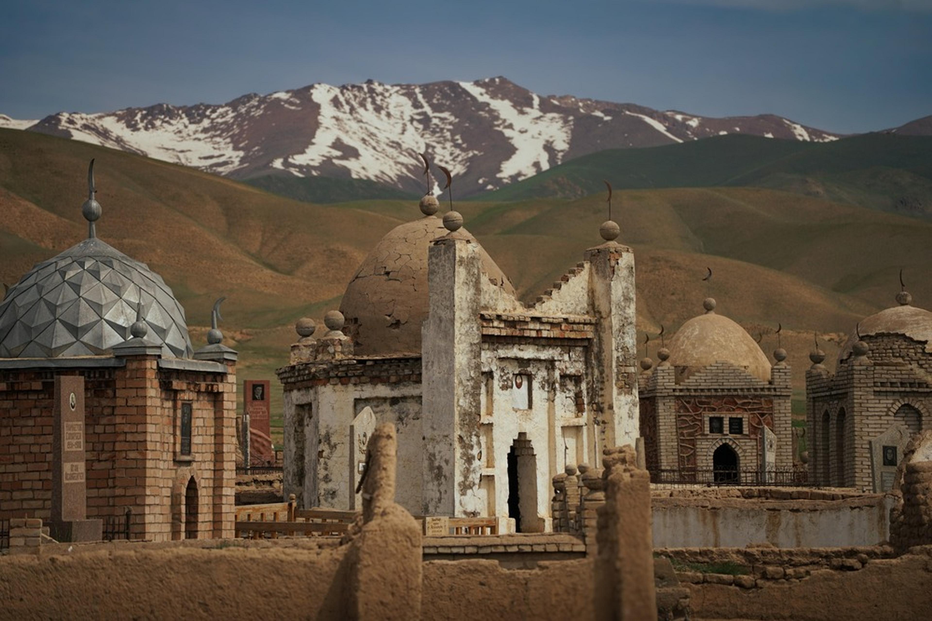 Ornate mausoleums with domed roofs stand against the backdrop of snow-capped mountains in a Kyrgyzstan landscape.