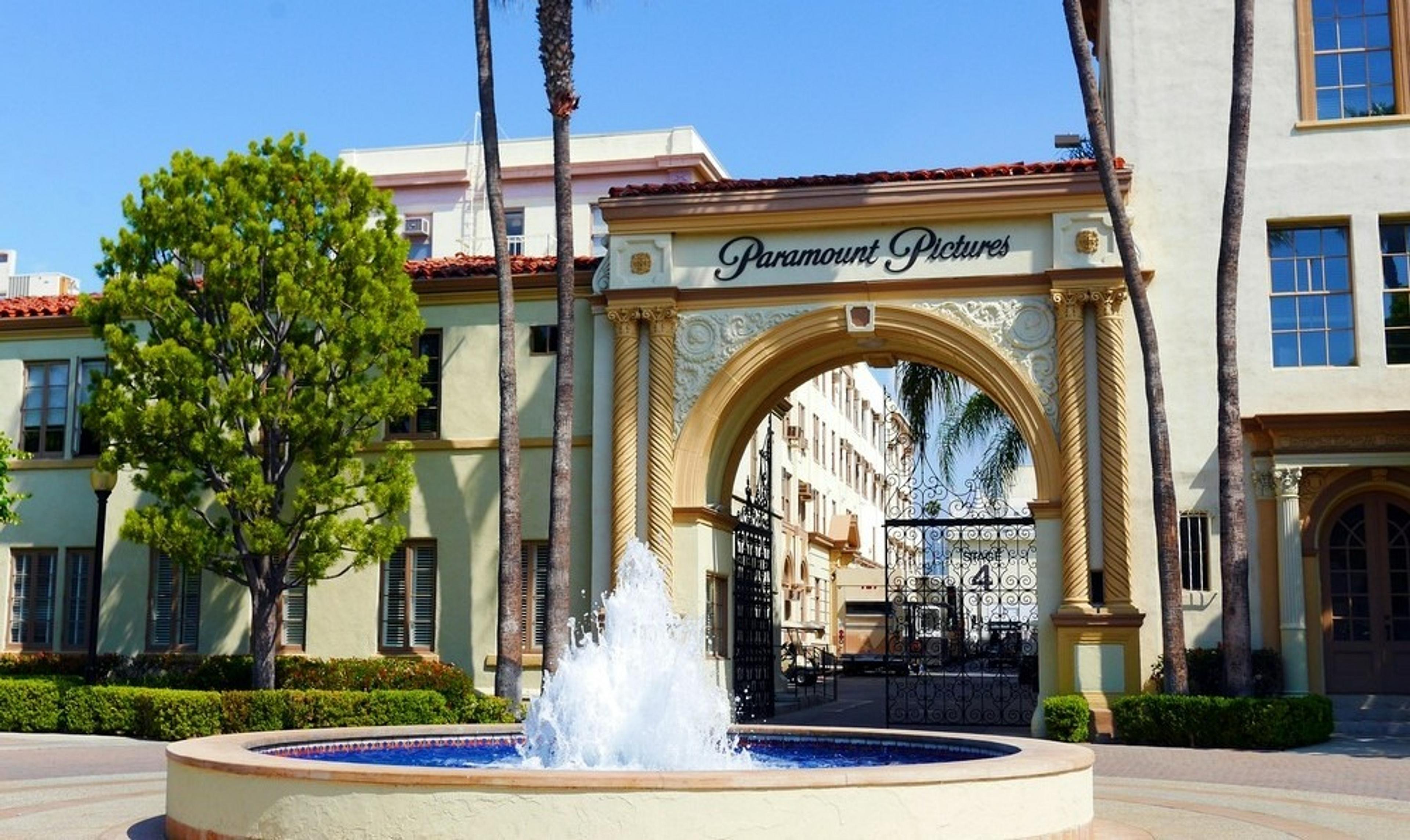 A grand archway labeled "Paramount Pictures" stands prominently at the entrance of the iconic film studio in Hollywood, California, surrounded by lush greenery and a fountain.