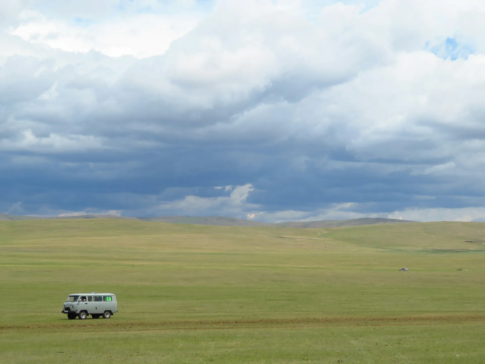 A van traverses the vast grasslands of the Mongolian steppe under a cloudy sky.