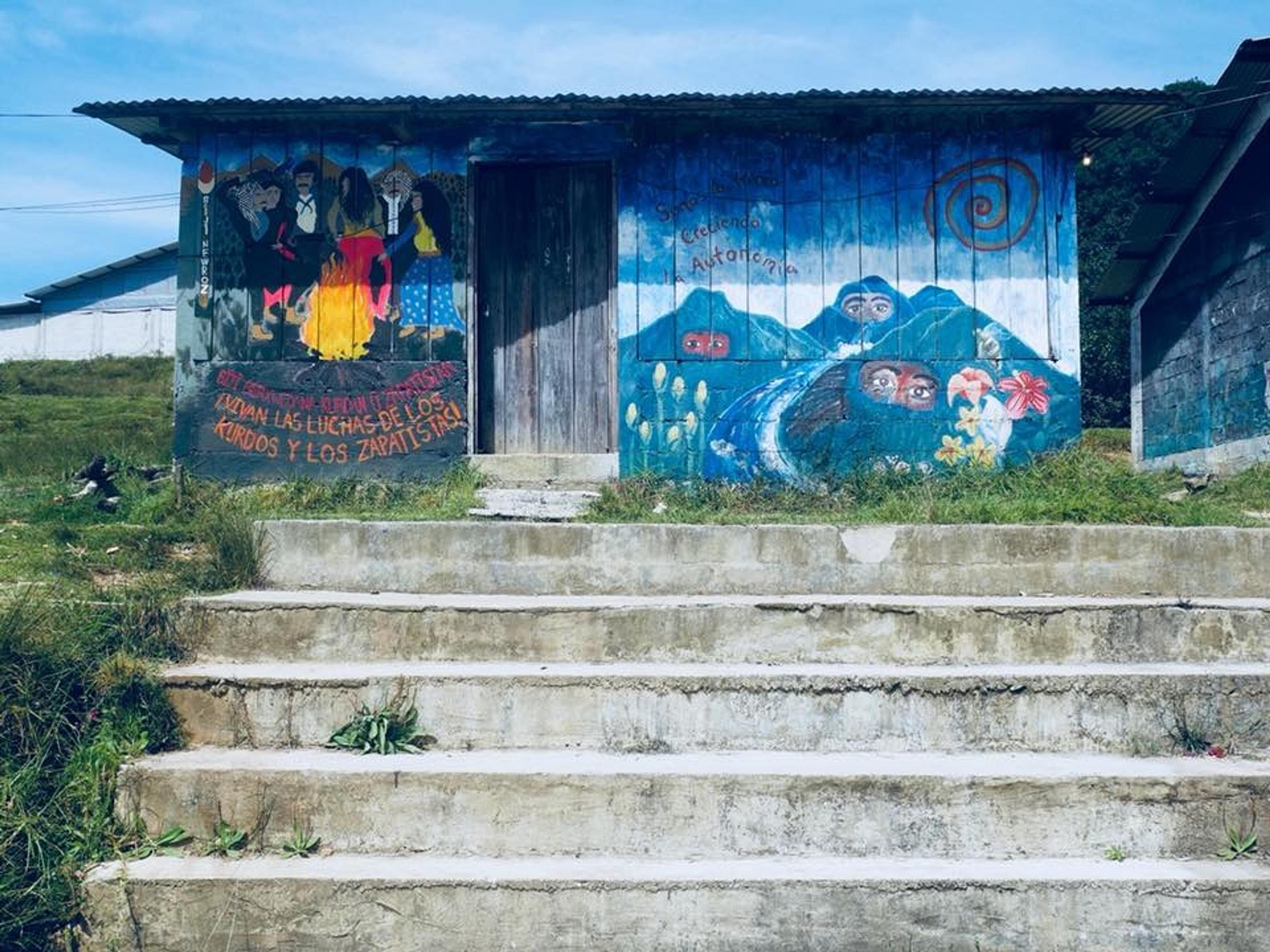 A vividly painted mural decorates a small building in a rural area of Chiapas, Mexico, featuring cultural and political motifs.