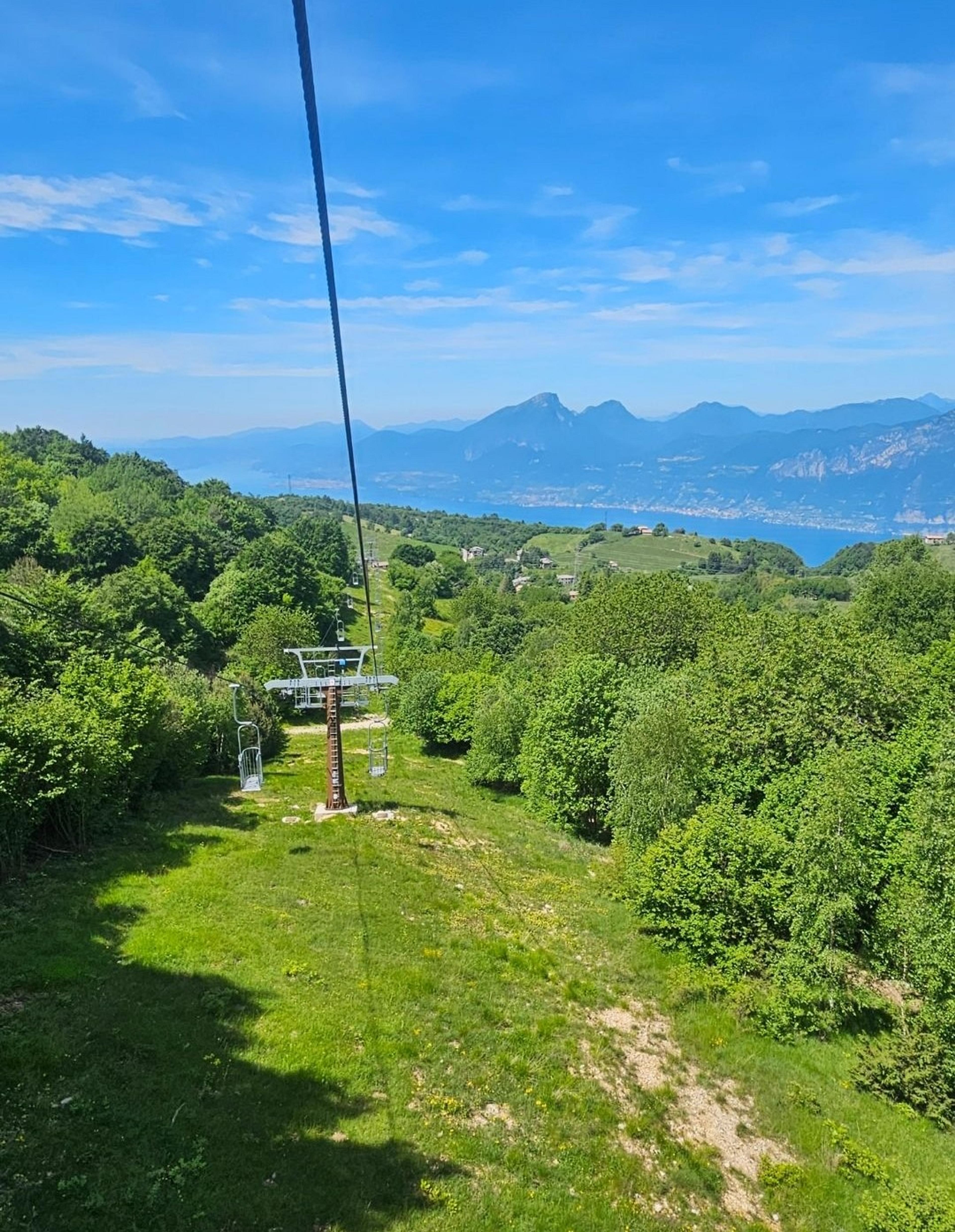 A scenic view overlooking Lake Garda in Italy, surrounded by lush greenery and mountains in the distance, with a cable car traversing the landscape.