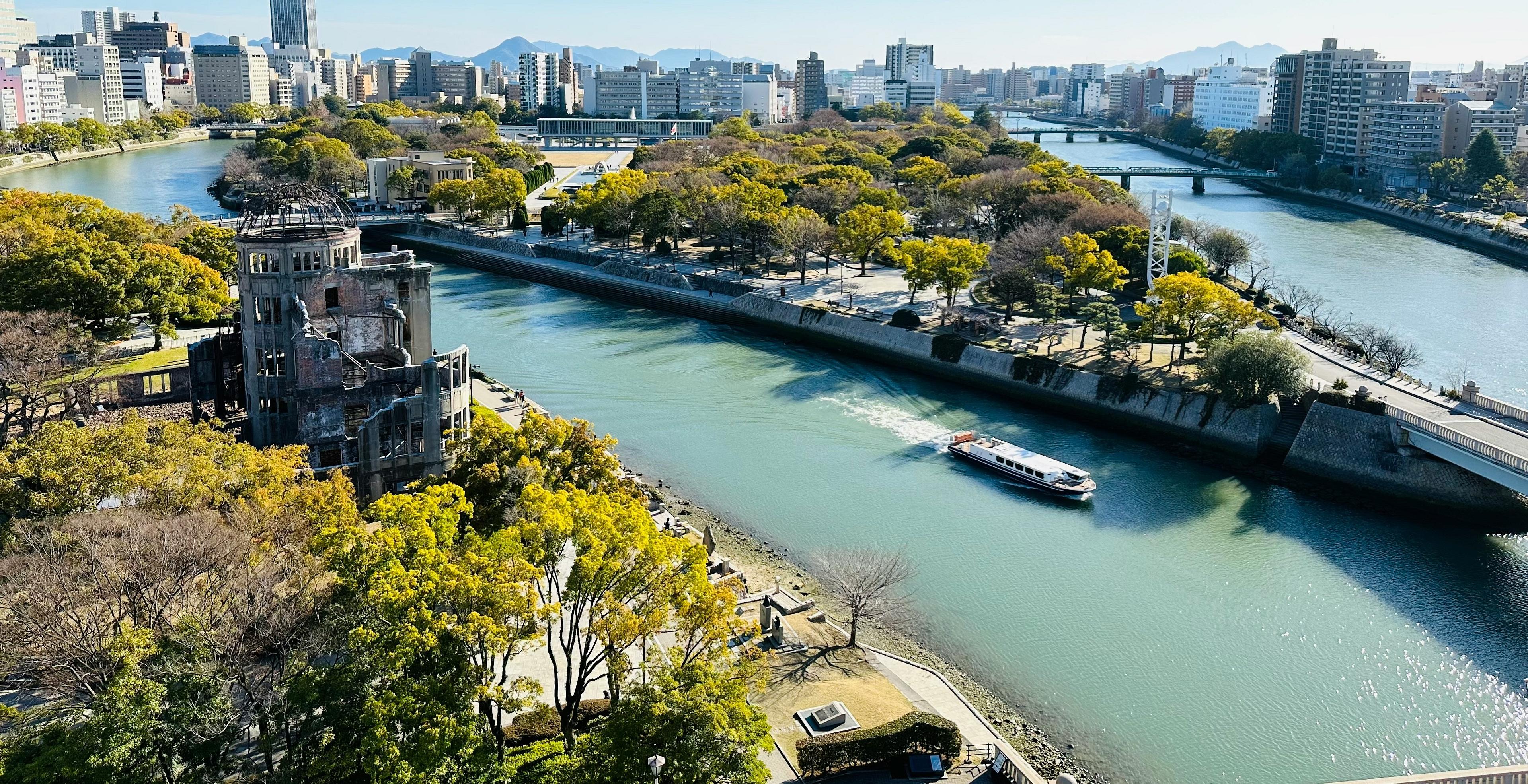 Hiroshima Peace Memorial (Atomic Bomb Dome) stands by a river surrounded by lush greenery and urban buildings in Hiroshima, Japan.