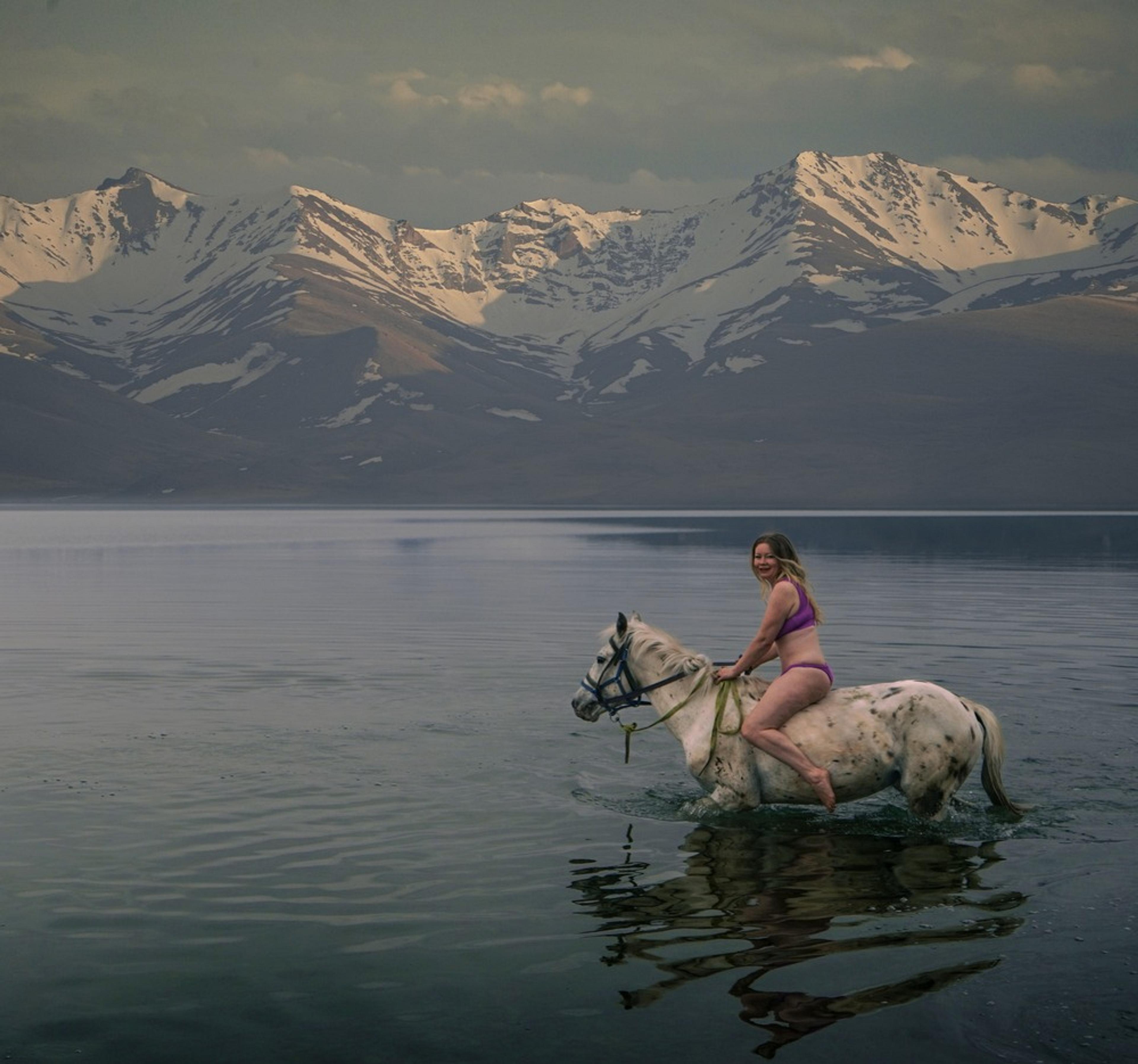 Flight Finder Kristi rides a white horse through the waters near the majestic, snow-capped mountains of Kyrgyzstan.