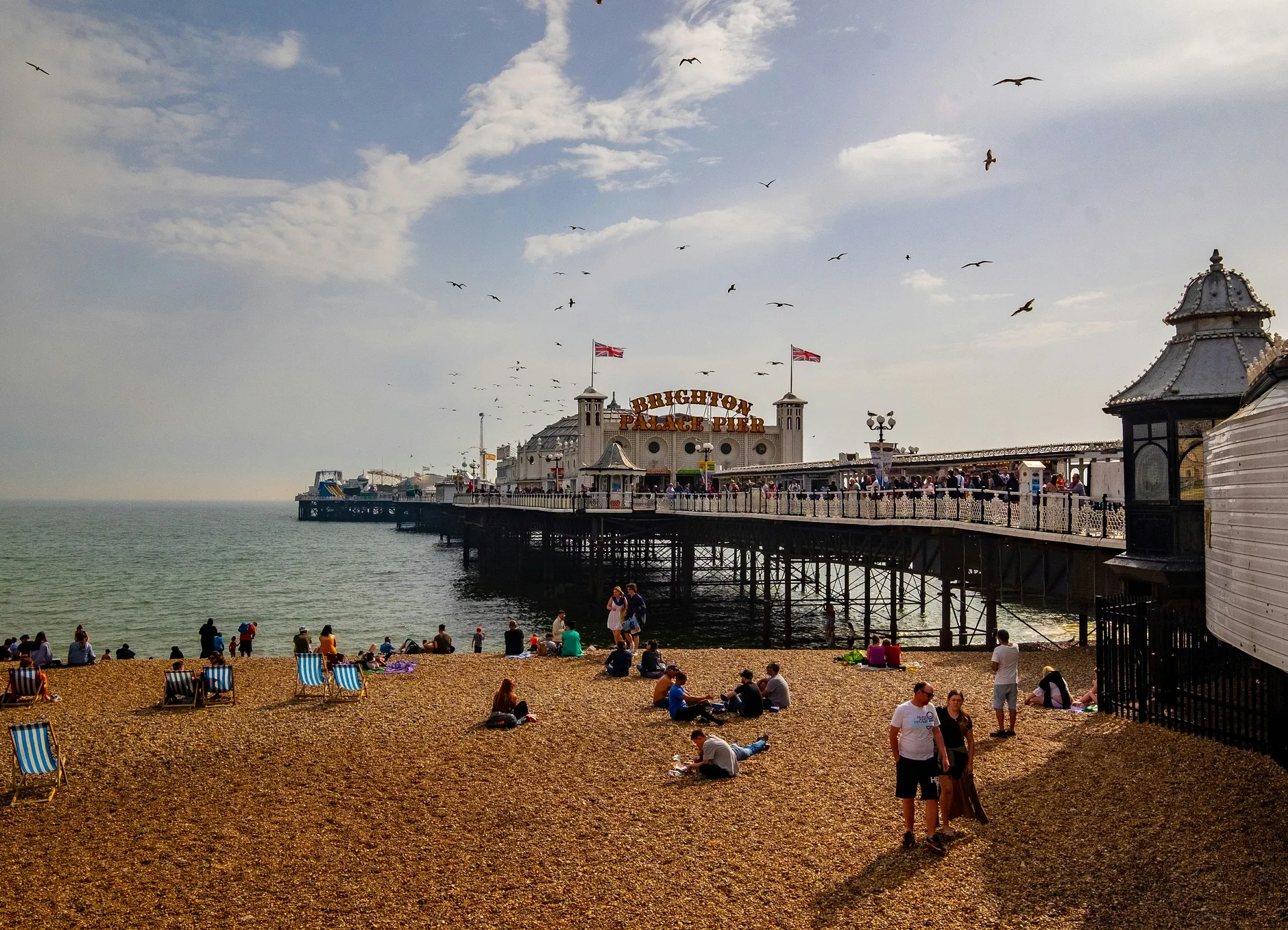 Brighton Palace Pier extends over the sea as people relax on the pebble beach beneath a sky filled with seagulls.