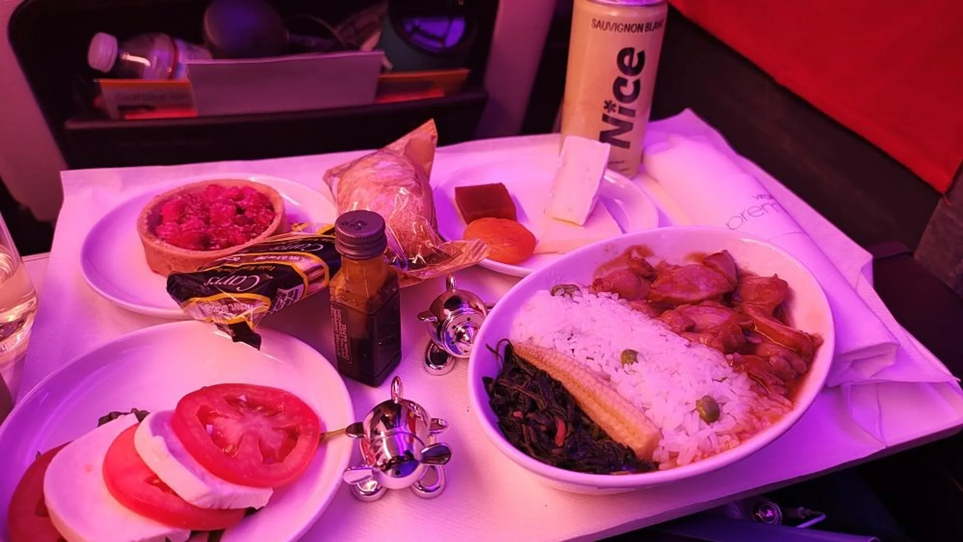 An upscale airplane meal with dishes like caprese salad, chicken curry with rice, and a raspberry tartlet is set on a tray inside a Virgin Atlantic cabin.