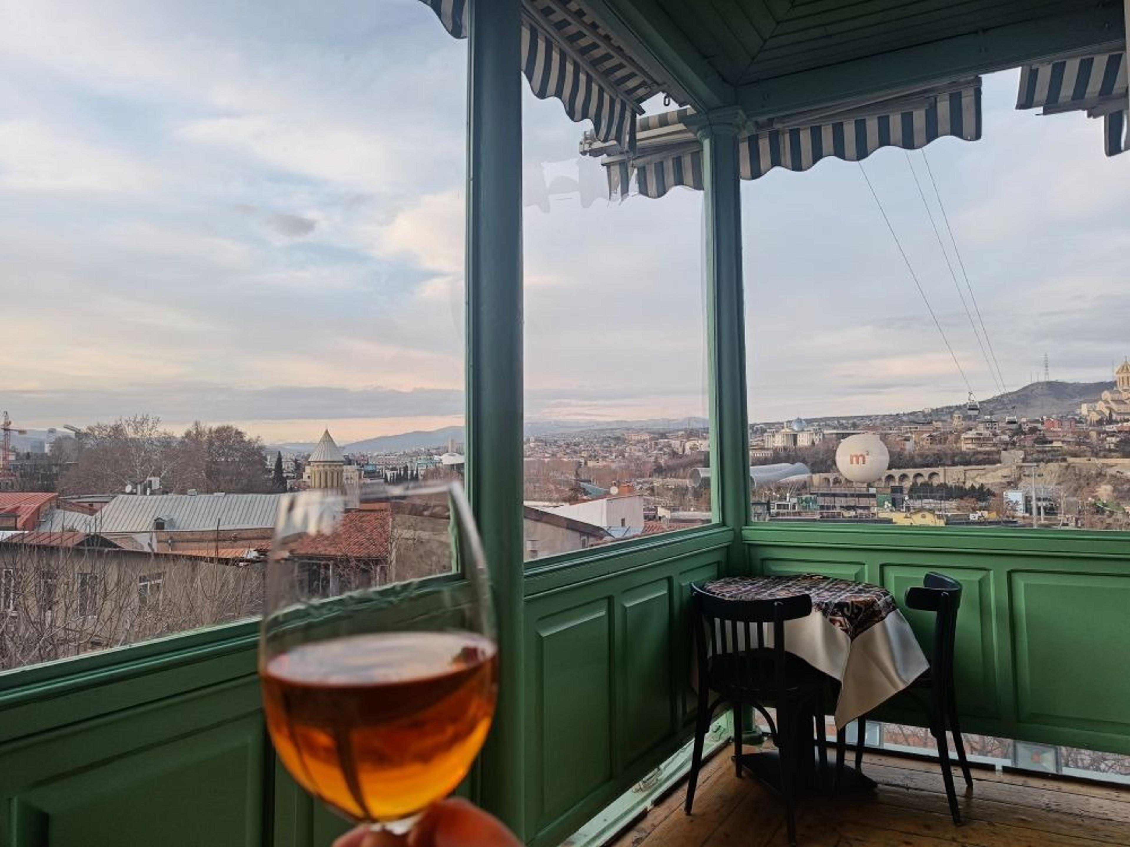 A picturesque view from a balcony in Tbilisi, Georgia, looking over the cityscape with a glass of wine in hand.