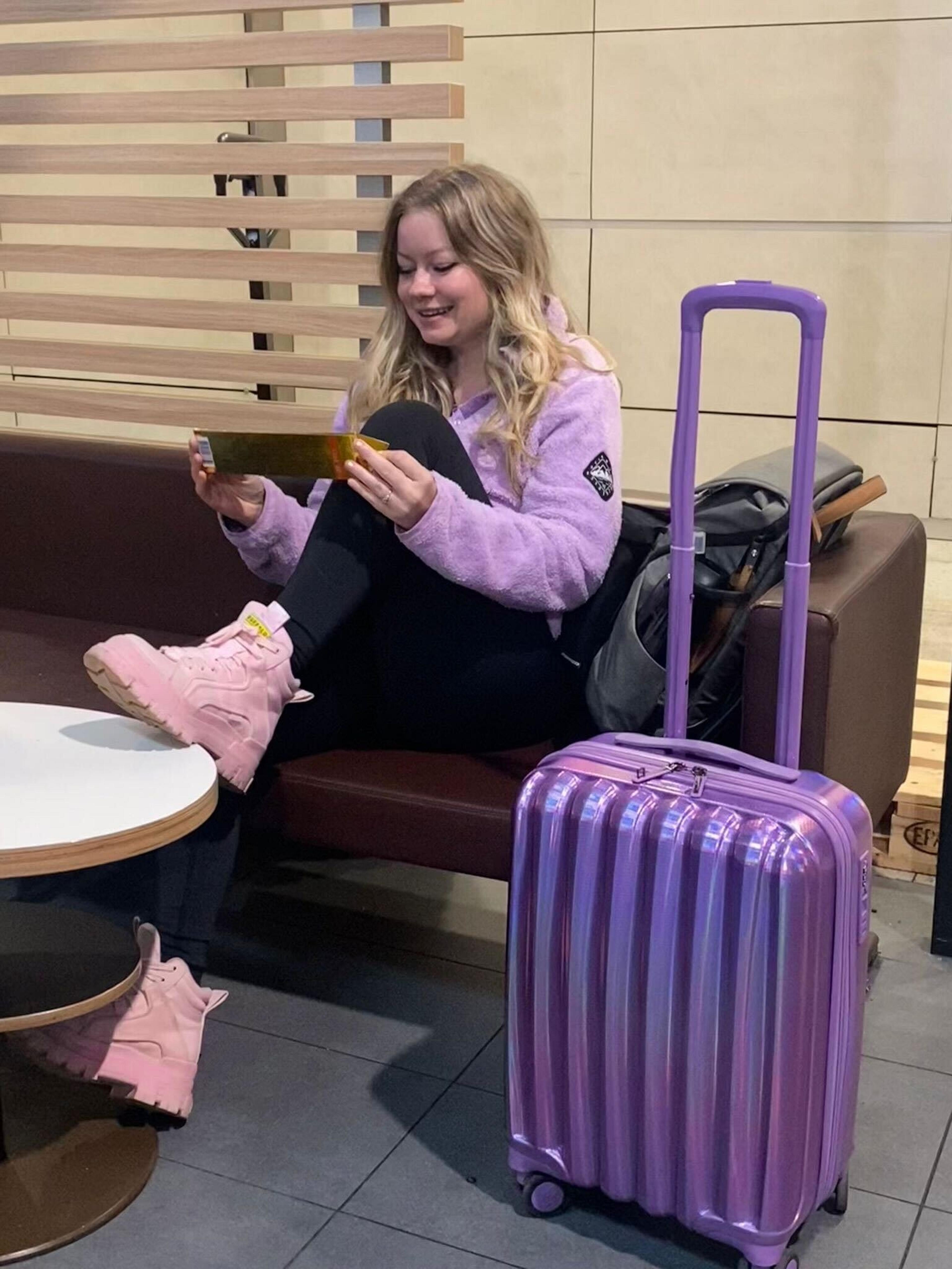 A traveler with a purple suitcase and pink boots relaxes in what appears to be an airport lounge.