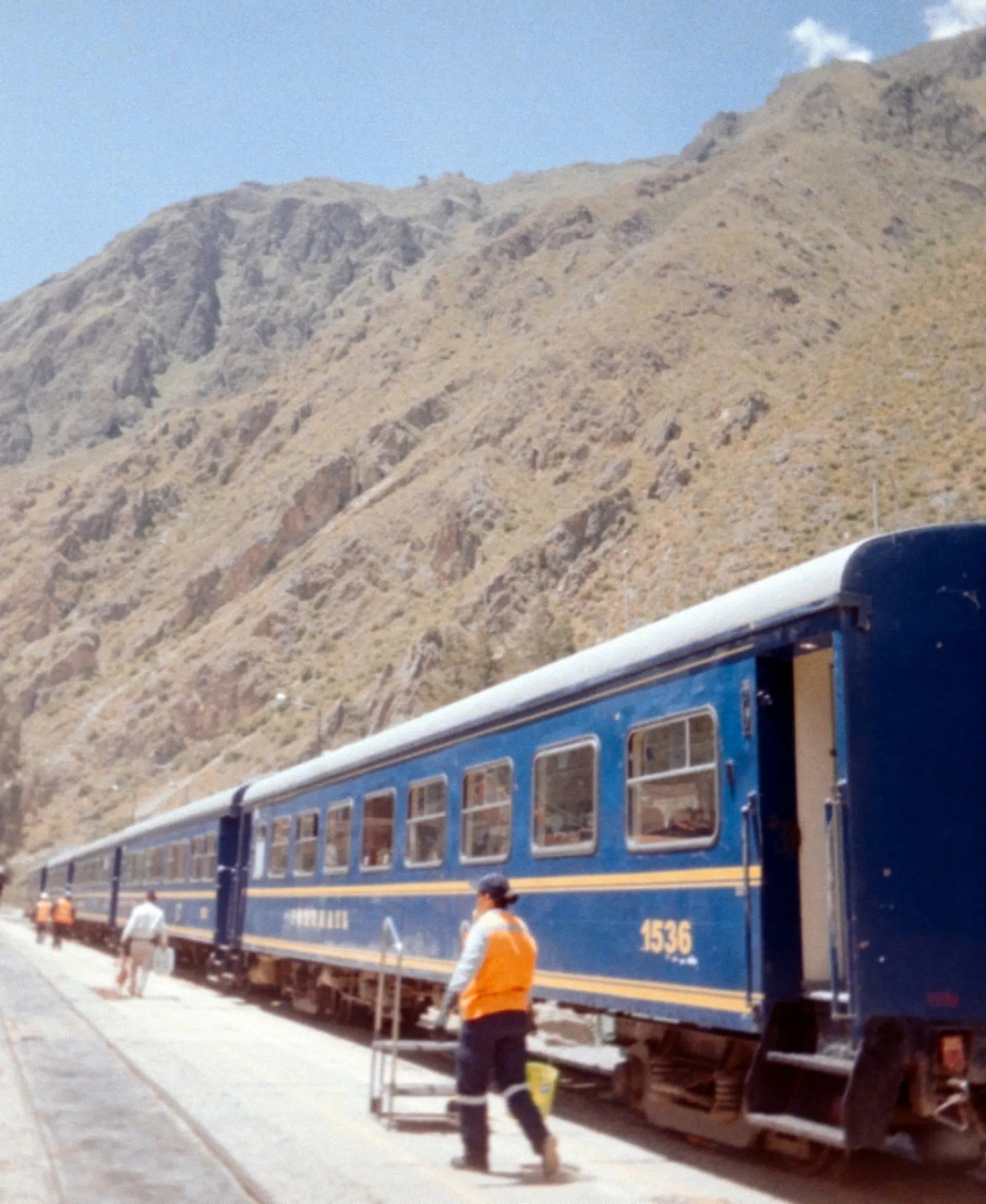 A blue train with yellow details stands on the tracks at the base of Peruvian mountains.