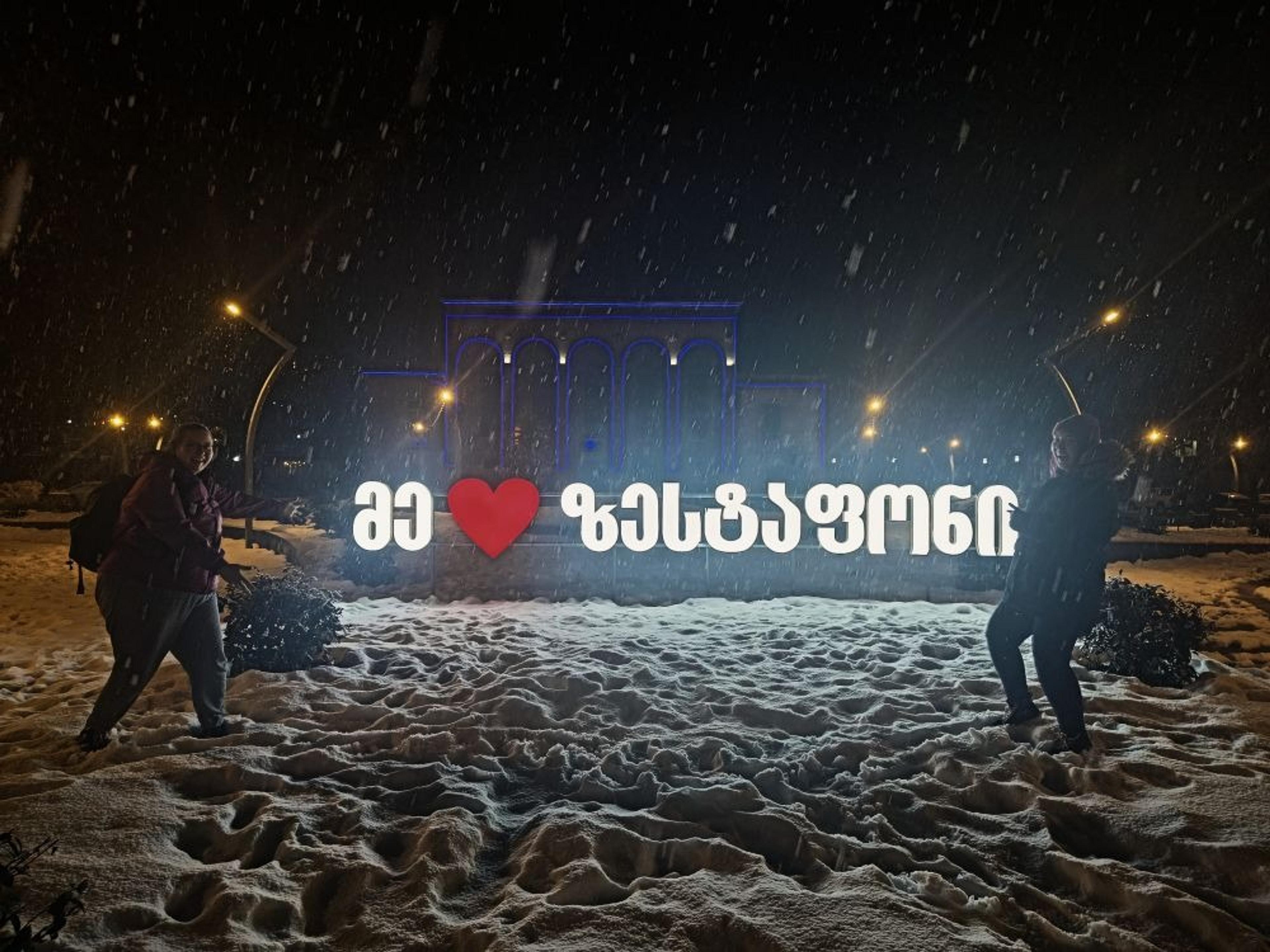 People stand in the snow at night in front of an illuminated sign in Zestafoni, Georgia.