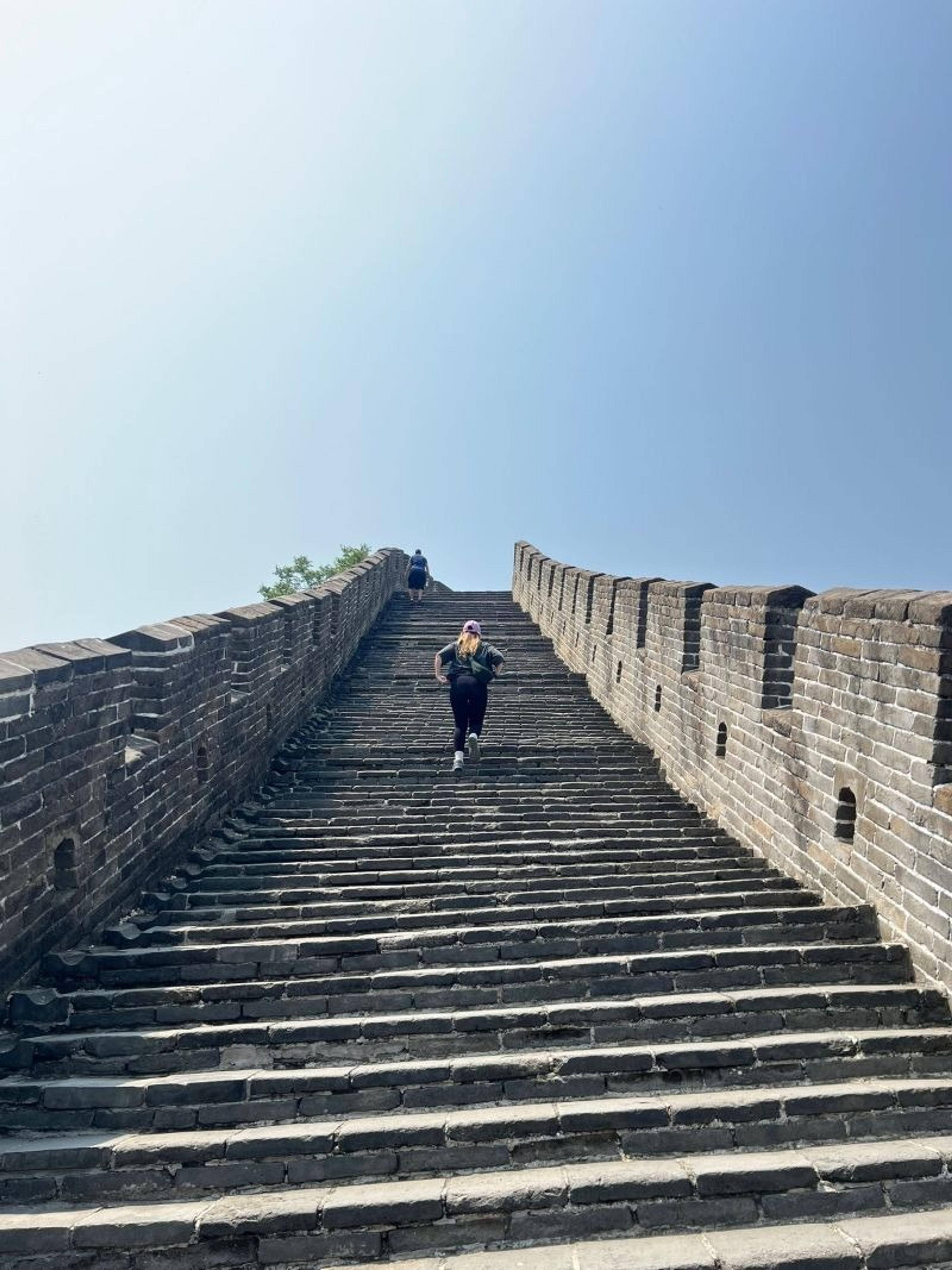 A person ascending a steep flight of steps on the Great Wall of China under a clear blue sky.