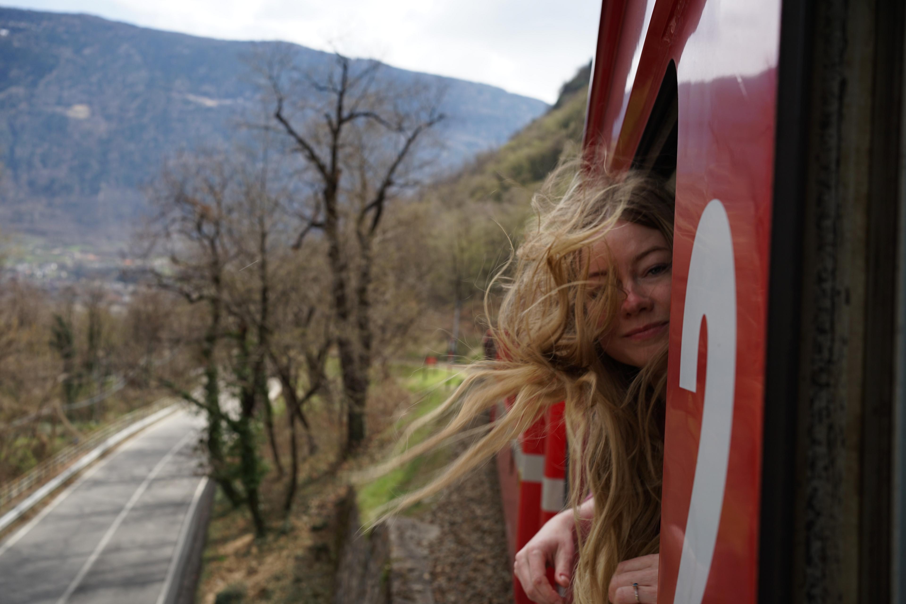 A woman leans out of a red train traveling through the scenic landscape of Valtellina in Italy, with mountains and a winding road in the background.ist