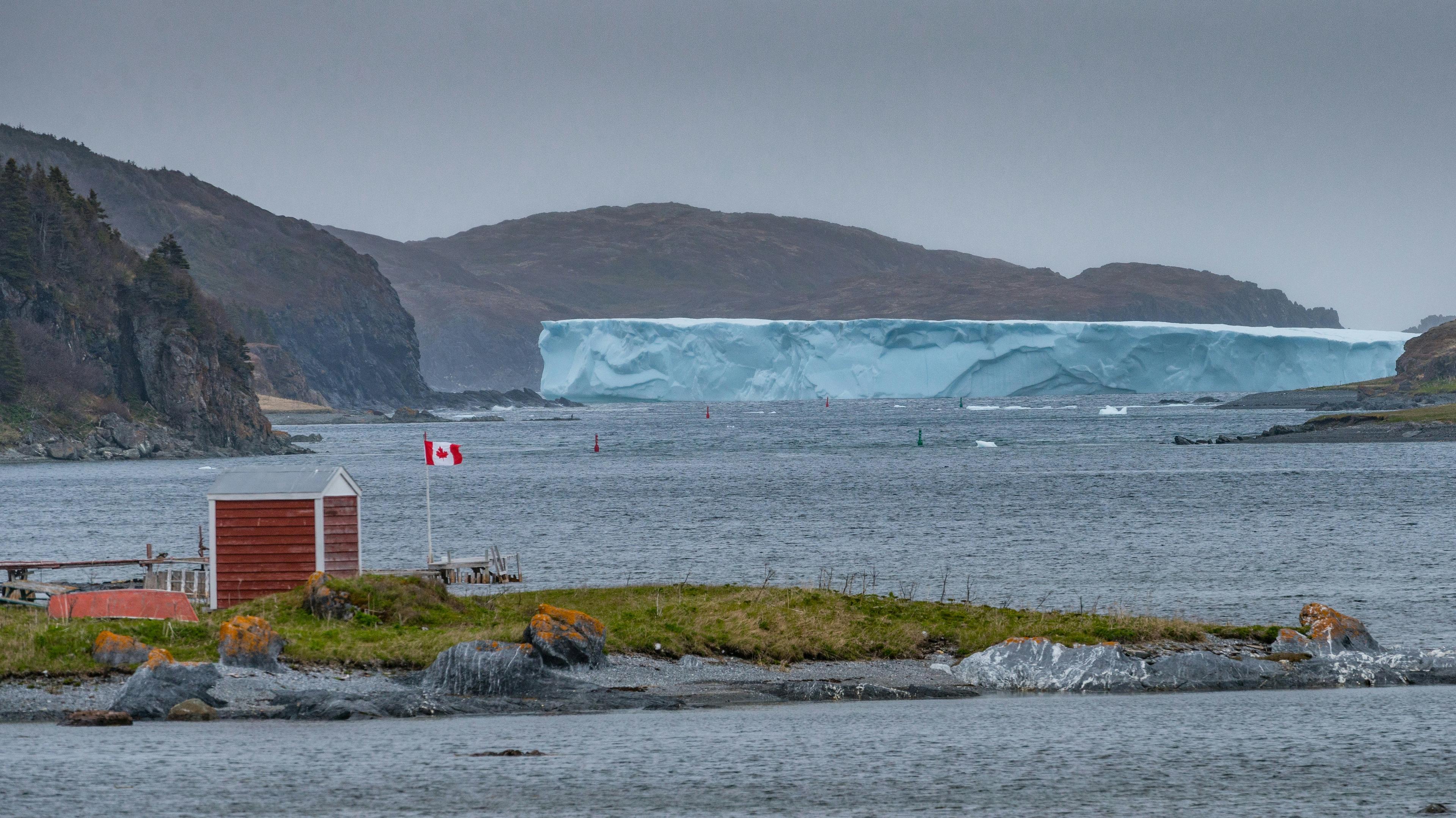 A massive iceberg drifts near Fogo Island, Newfoundland, with a Canadian flag flying beside a small red building by the shore.