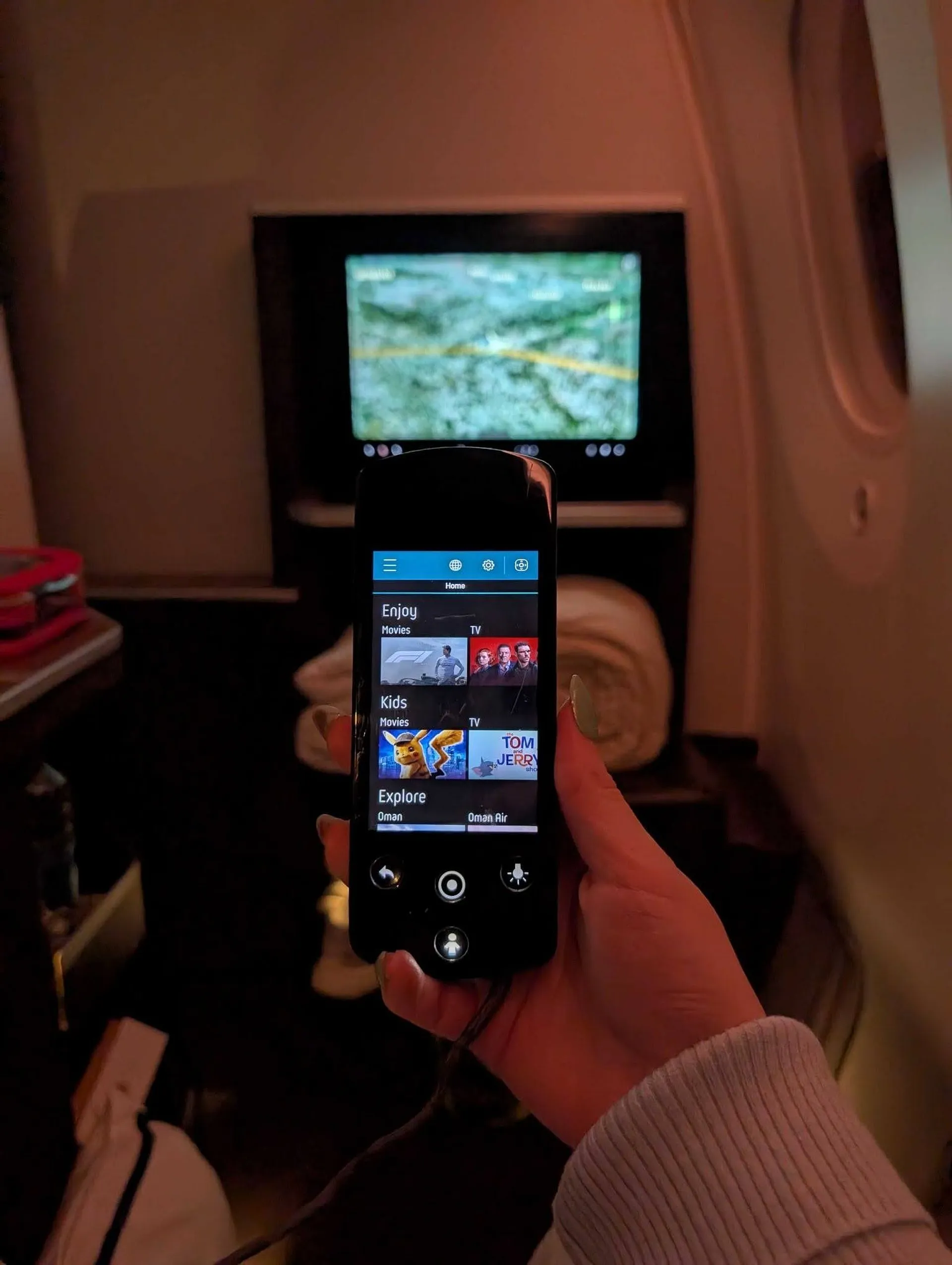 A passenger on an Oman Air flight browses the in-flight entertainment options on a remote control with a map displayed on the personal screen.