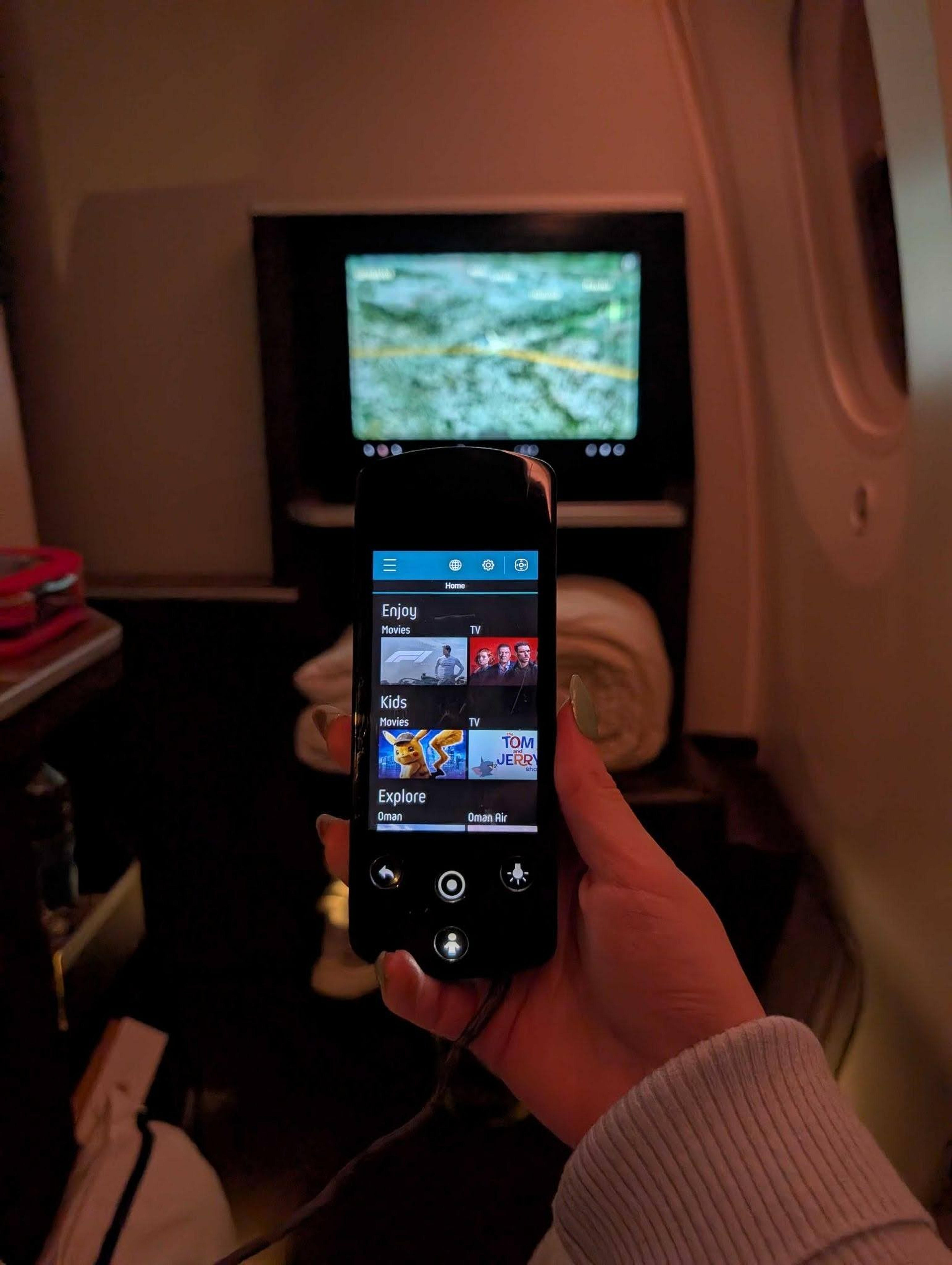 A passenger on an Oman Air flight browses the in-flight entertainment options on a remote control with a map displayed on the personal screen.