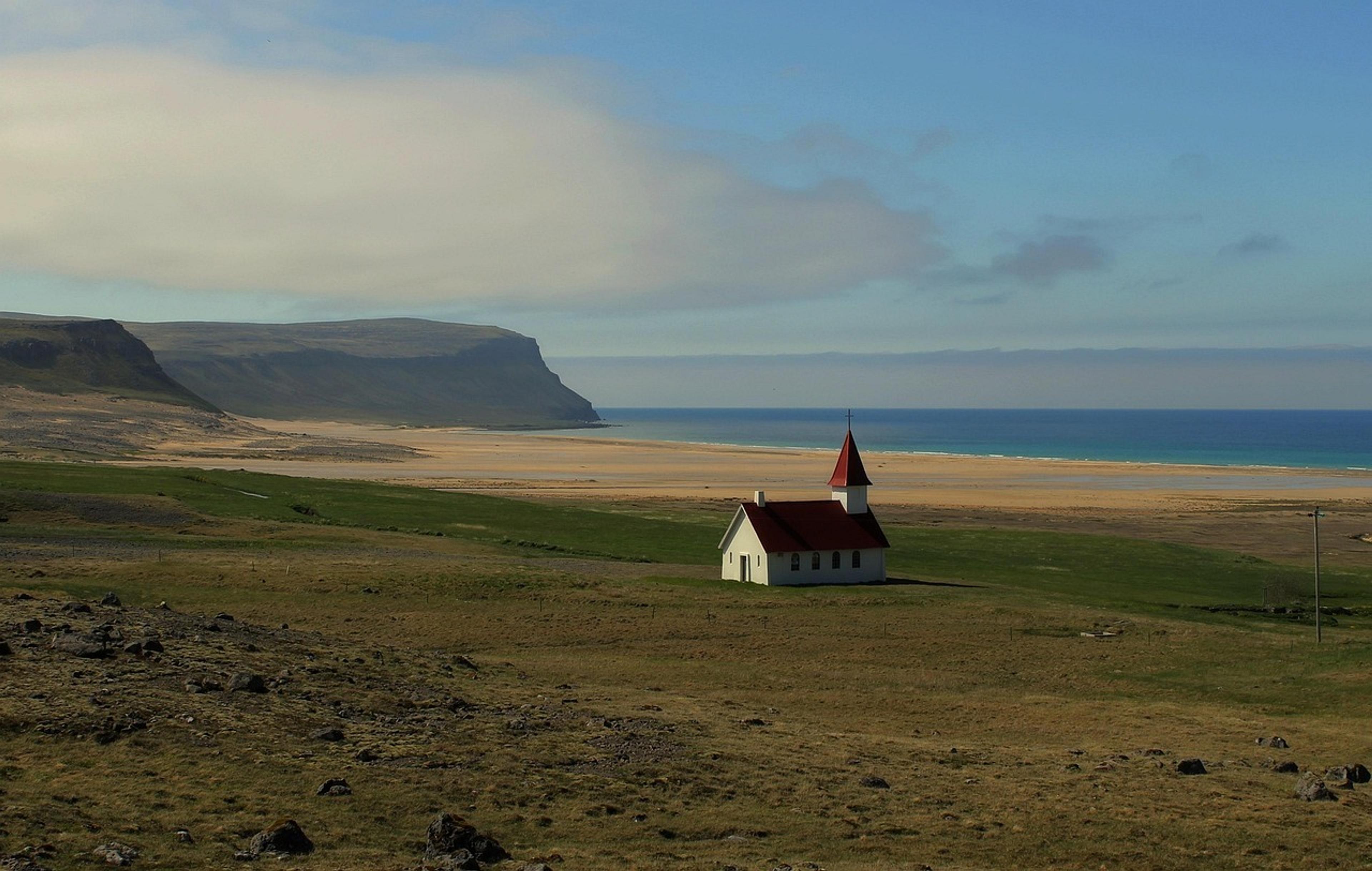 A solitary red-roofed church stands near a vast sandy beach in the Westfjords region of Iceland.
