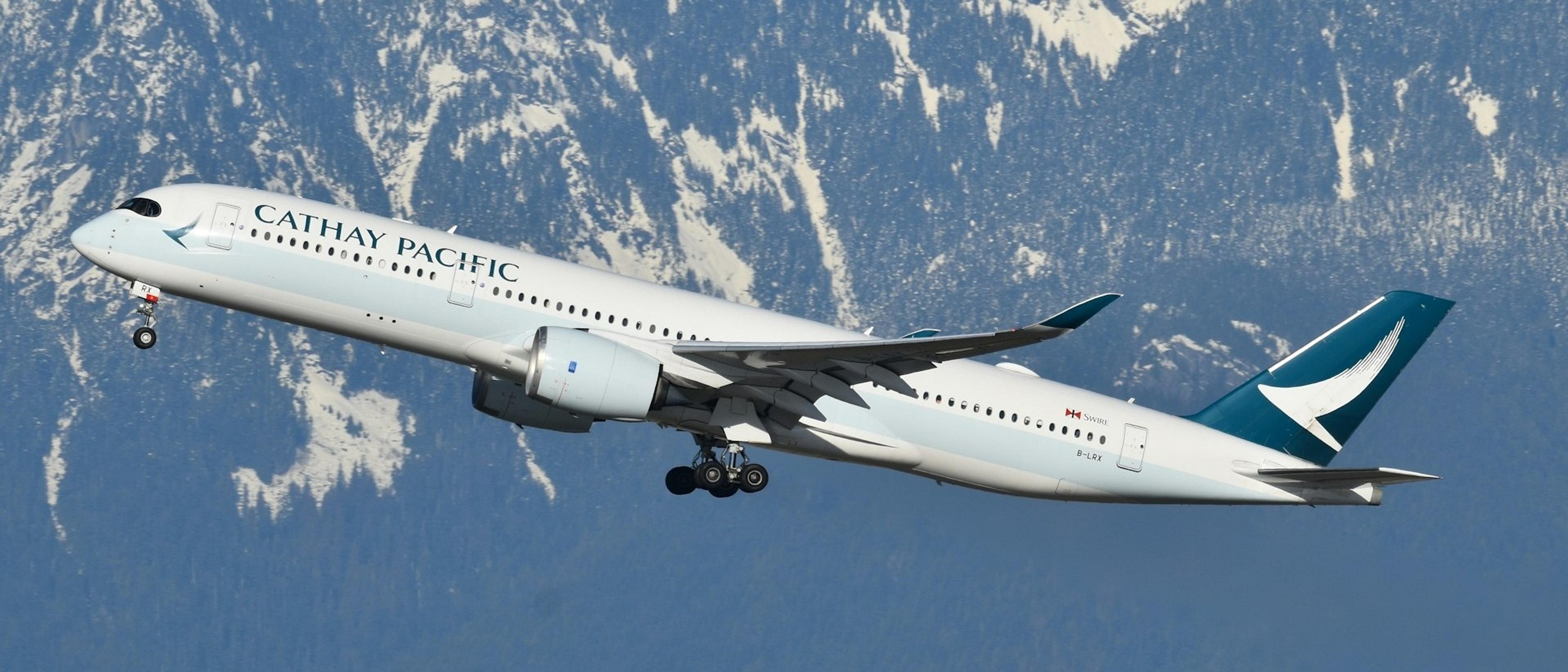 A Cathay Pacific airplane ascends against the backdrop of snow-capped mountains, likely over British Columbia, Canada.