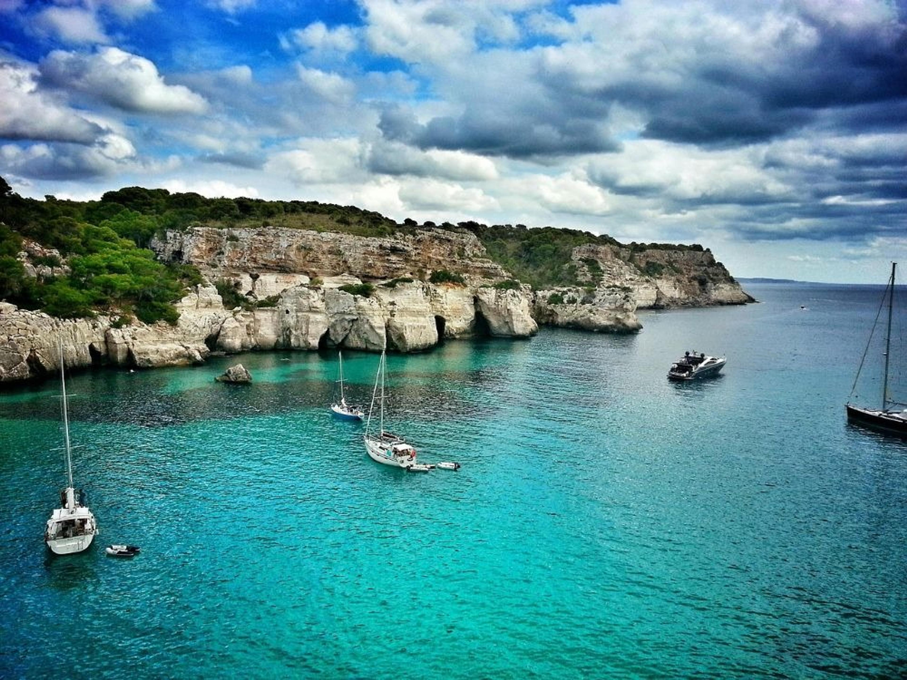 Sailboats and cliffs grace the turquoise waters of Cala Macarella in Menorca, Spain.