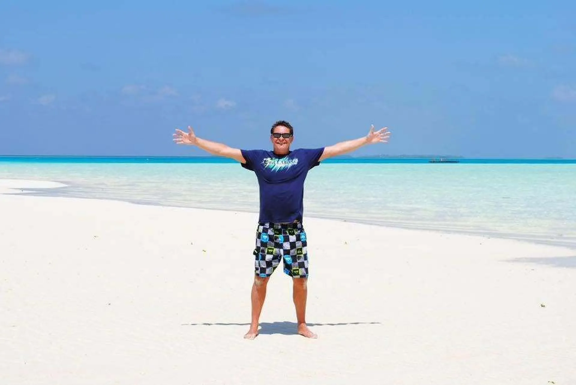 A person stands gleefully on a pristine white-sand beach with crystal-clear turquoise waters in the Maldives.