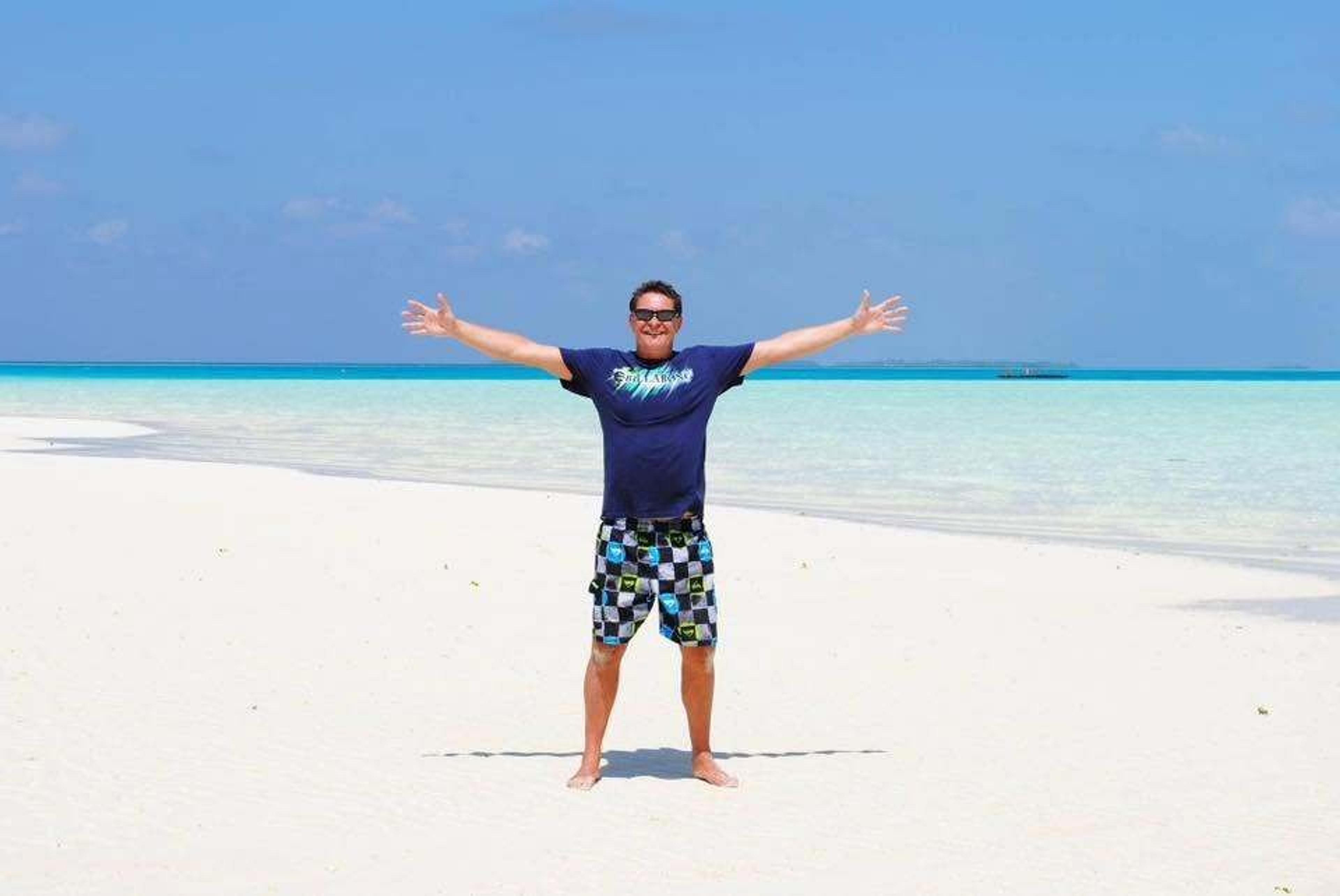 A person stands gleefully on a pristine white-sand beach with crystal-clear turquoise waters in the Maldives.