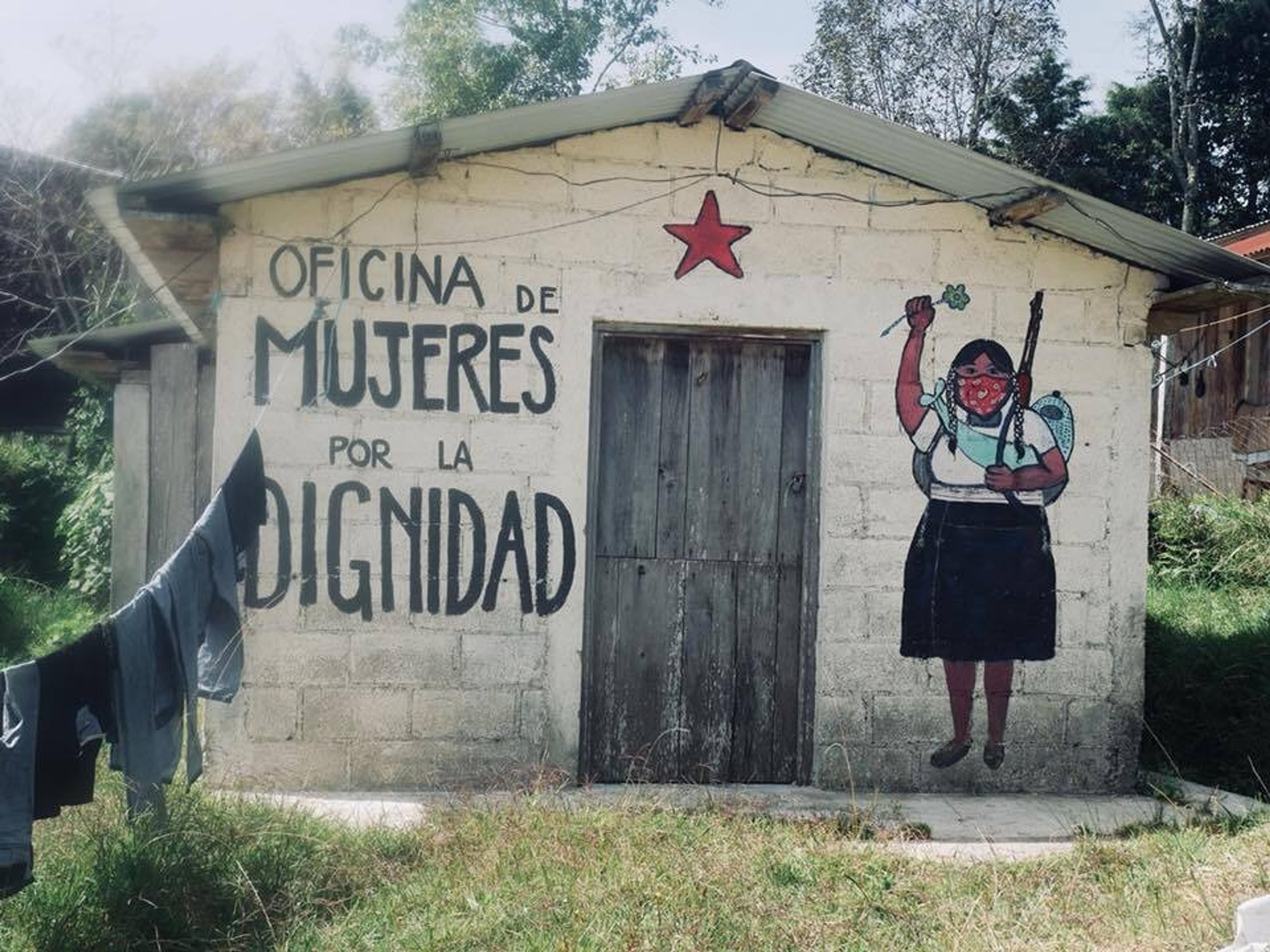 A mural with the words "Oficina de Mujeres por la Dignidad" decorates a small building in Chiapas, Mexico, featuring an empowering image of a woman holding a rifle and a flower.