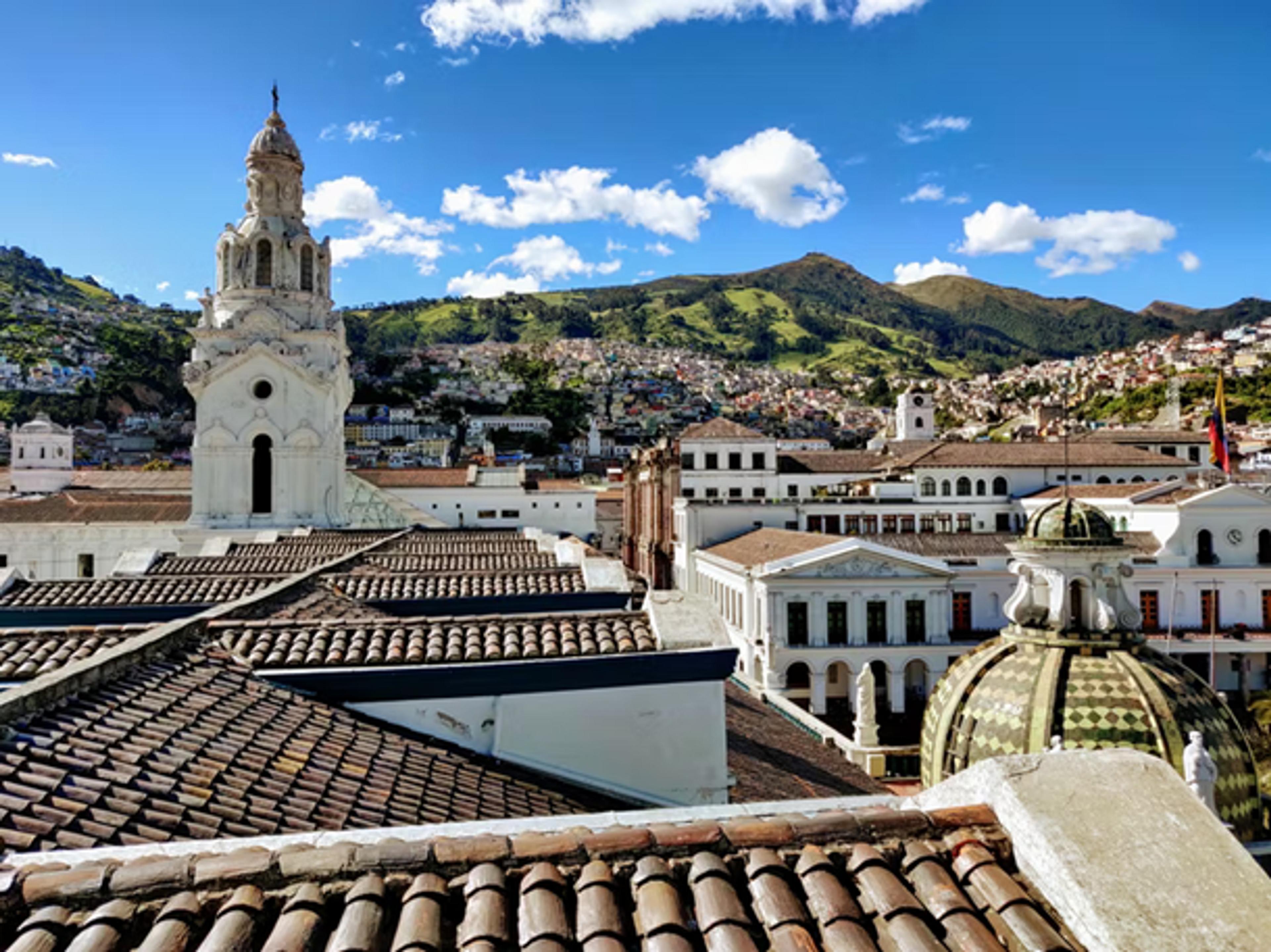 A scenic view of Quito, Ecuador, featuring colonial architecture with mountains in the background under a bright blue sky.